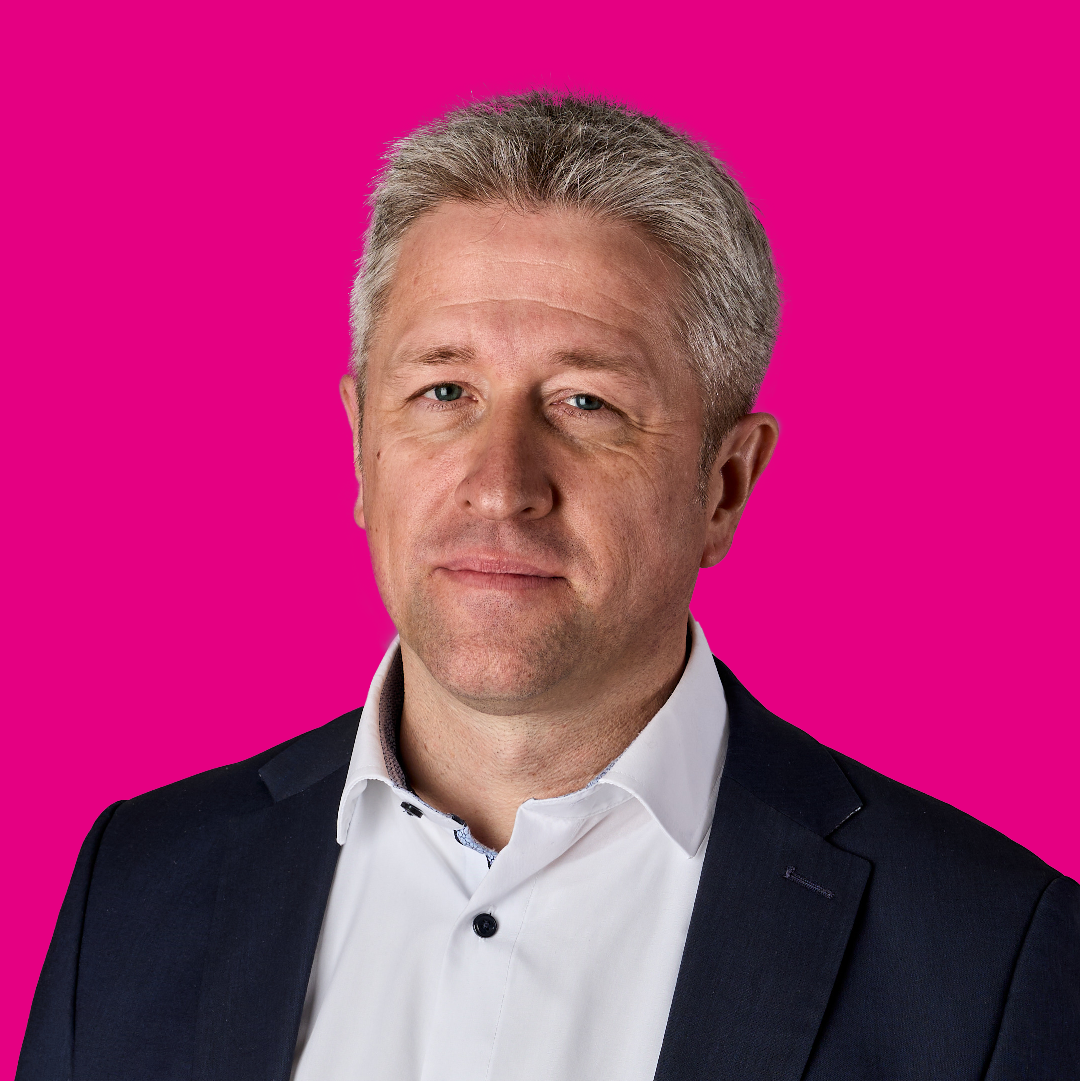Porträt eines Mannes mit grauem Haar in formeller schwarzer Jacke und weißem Hemd vor pinkem Hintergrund.
