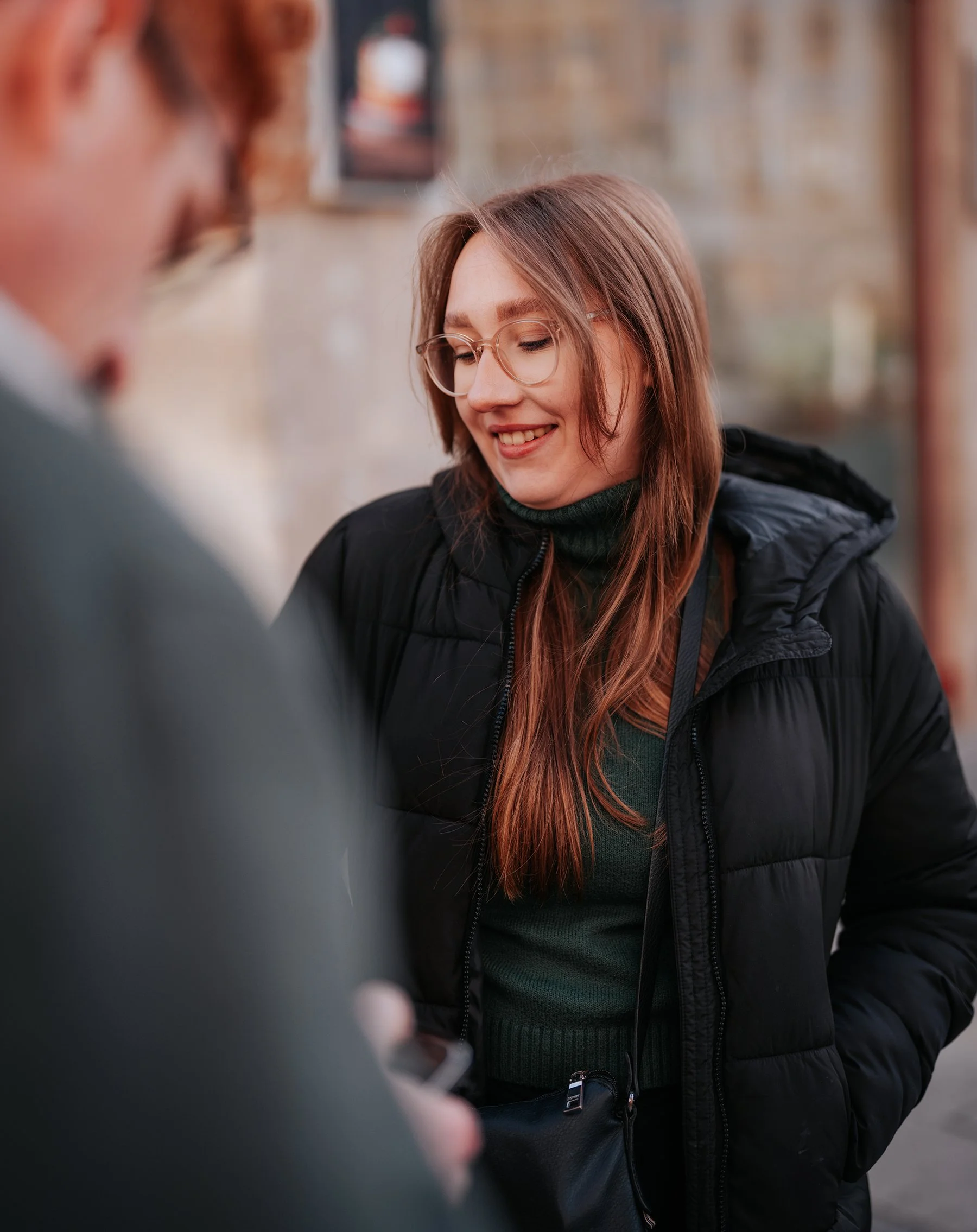 Eine Frau mit Brille, braunen Haaren und dunkler Jacke lächelt. Im Vordergrund ist eine Person mit grauem Mantel und Handy, unscharf.
