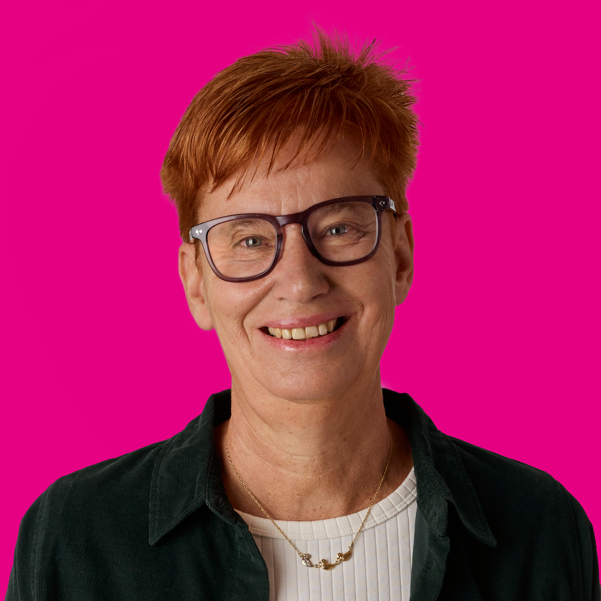 Eine lachende Frau mit roten Haaren, Brille und schwarzer Jacke vor einem pinken Hintergrund.