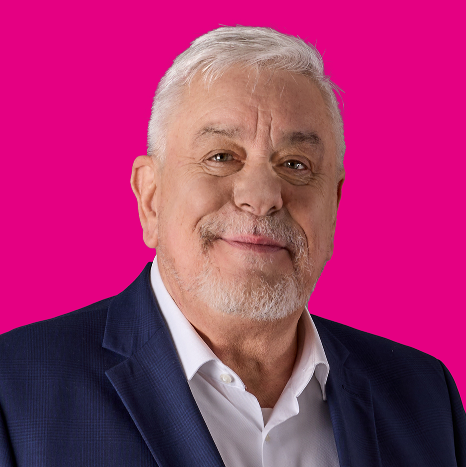 Porträt eines älteren Mannes mit weißen Haaren und Bart, trägt einen dunkelblauen Anzug und ein weißes Hemd, vor pinkem Hintergrund.