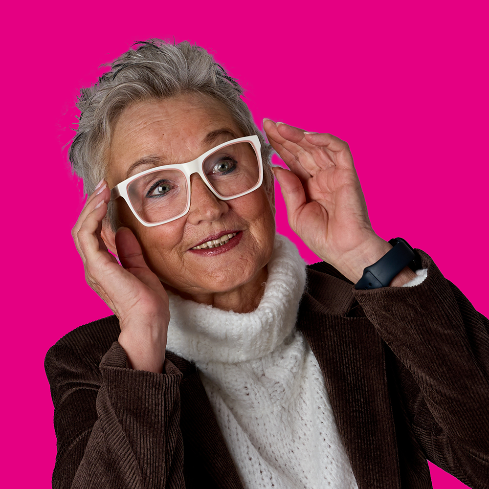Ältere Frau mit kurzen grauen Haaren, trägt große weiße Brille, weiße Pullover und braunen Cordjacke, positiver Gesichtsausdruck, steht vor pinkem Hintergrund.