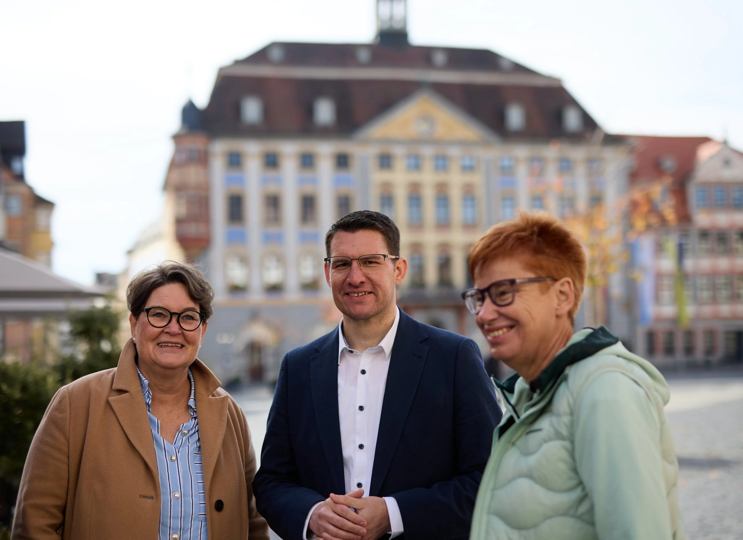 Drei Menschen, zwei Frauen und ein Mann, stehen im Freien vor einem historischen Gebäude, alle lächeln.