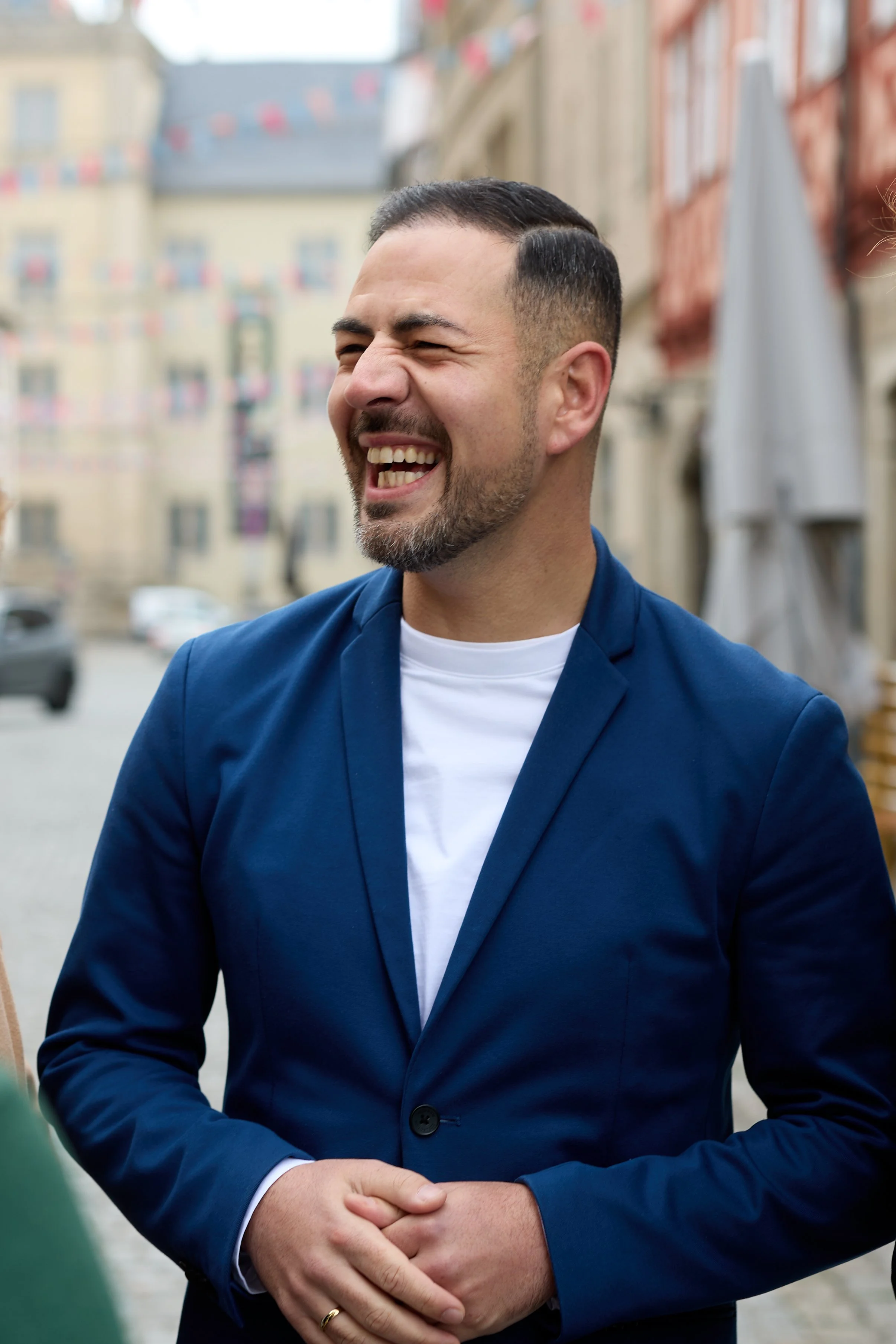 Ein lachender Mann in einem blauen Anzug und weißen T-Shirt steht draußen, vermutlich auf einer Straße in einer Stadt.