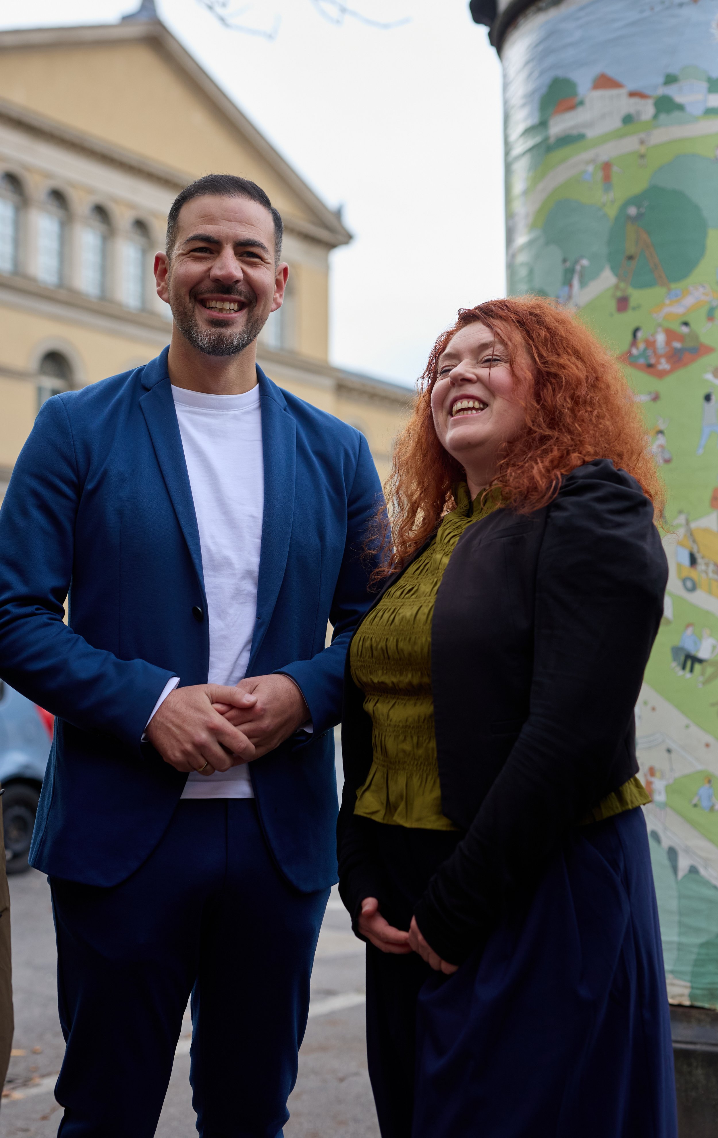 Zwei lachende Menschen, ein Mann in blauemAnzug und eine Frau, stehen draußen vor einem Mural mit einer grünen Landschaft und vielen Menschen beim Spielen, bei einem öffentlichen Ereignis.
