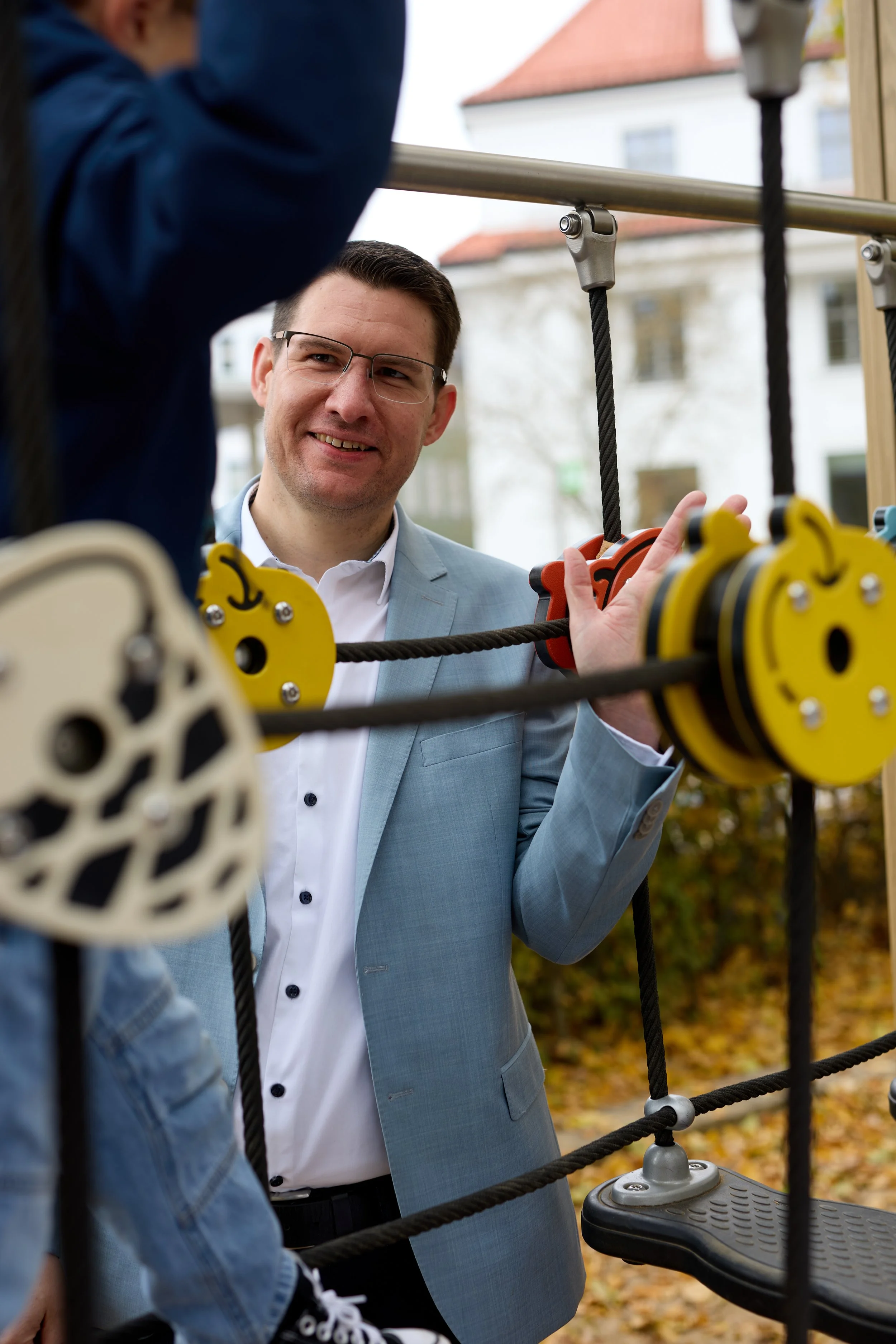 Ein Mann in einem grauen Anzug unterhält sich mit einem Kind auf einer Kletterpyramide im Freien, im Hintergrund sind Häuser und herbstliche Bäume.