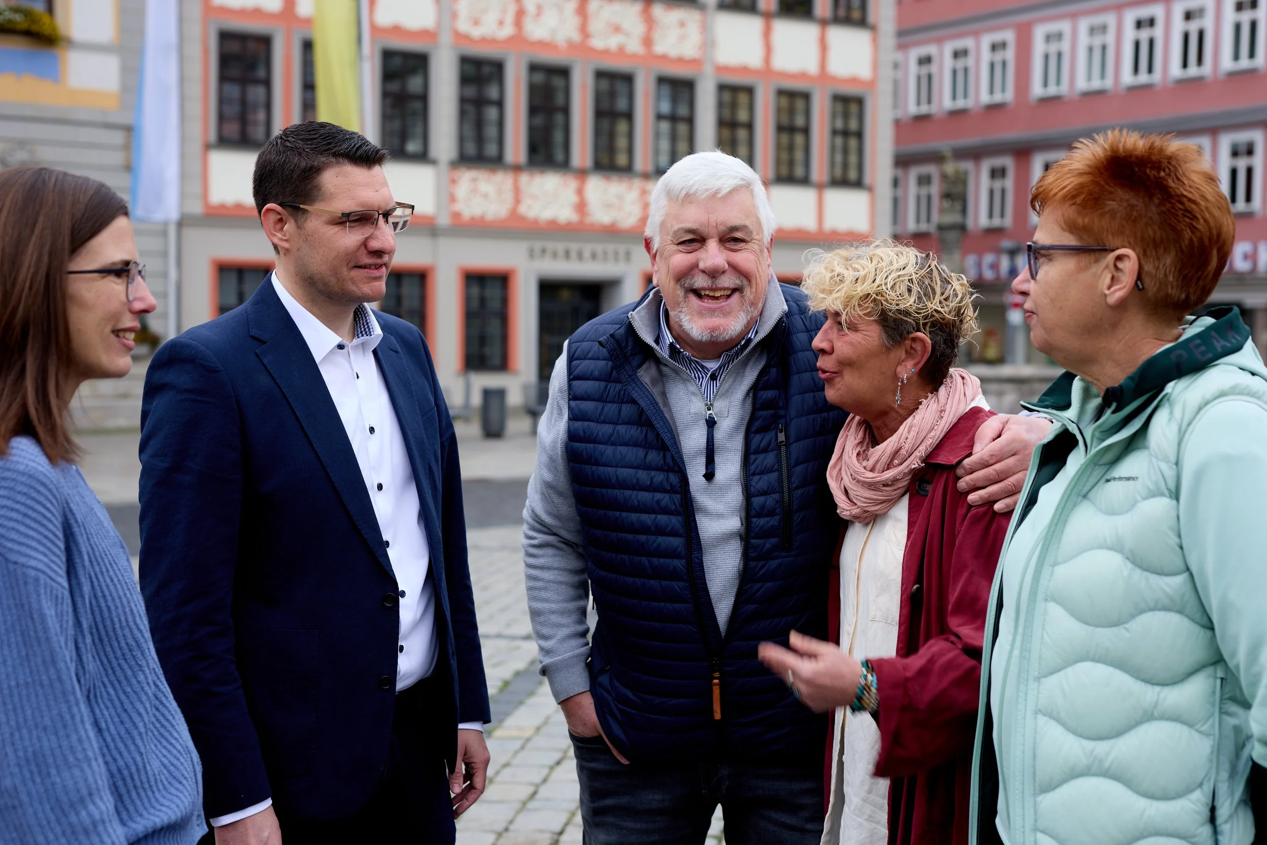 Fünf Personen lachen und sprechen draußen vor einem bunten Gebäude.