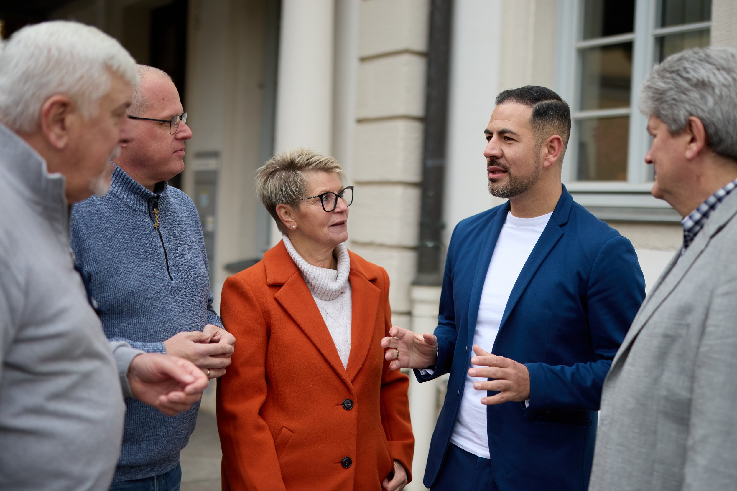 Eine Gruppe von fünf Menschen im Gespräch vor einem Gebäude, es wirkt wie eine Diskussion oder ein Austausch.
