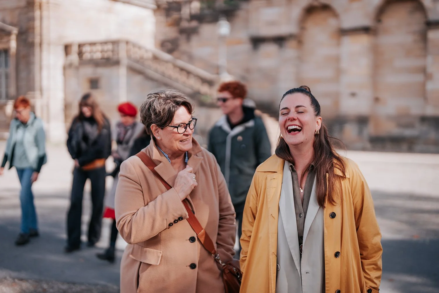 Zwei lachende Frauen im Gespräch auf einer Straße mit historischen Gebäuden im Hintergrund, im Hintergrund weitere Menschen.