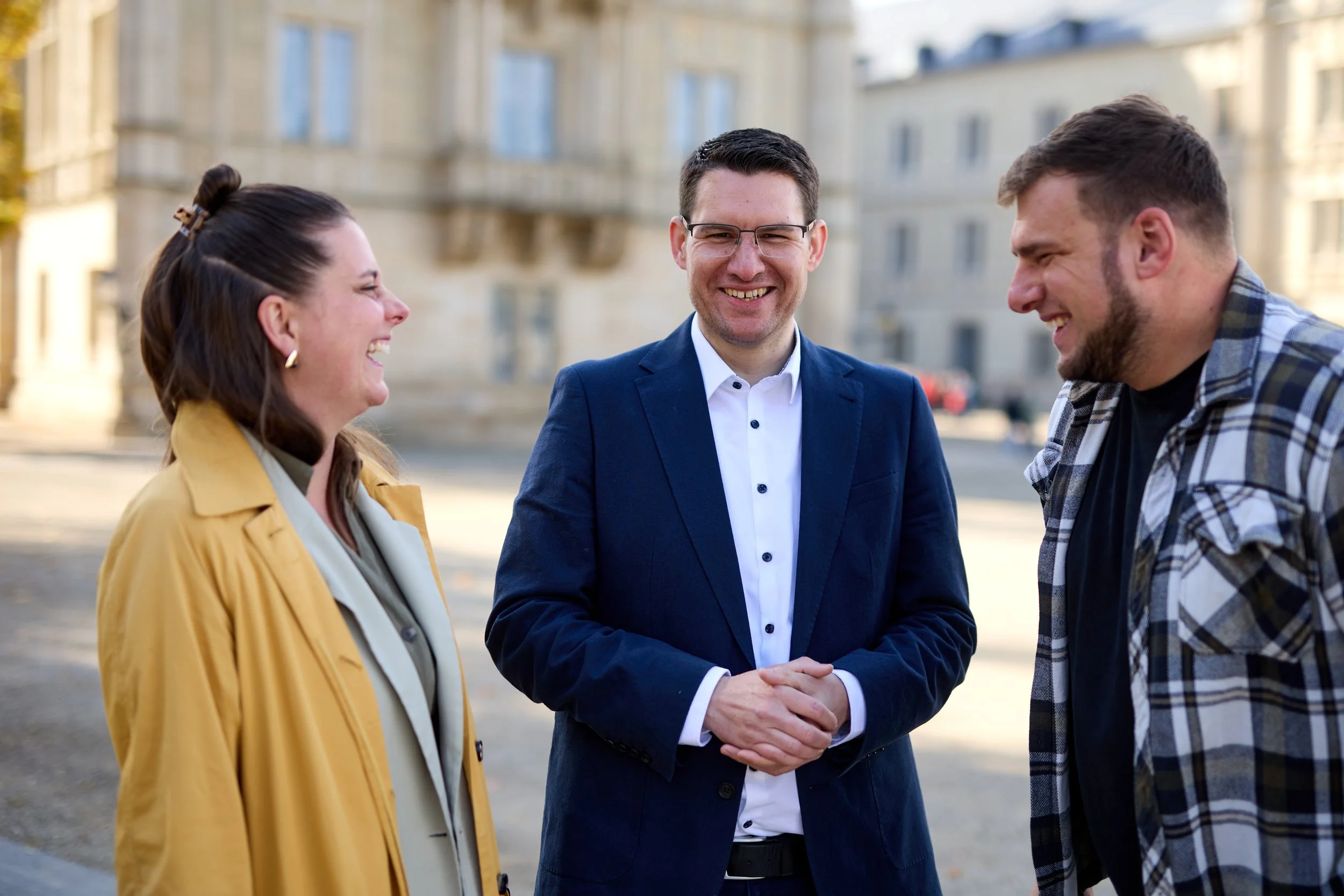 Drei Personen, zwei Männer und eine Frau, unterhalten sich lachend im Freien vor einem historischen Gebäude.