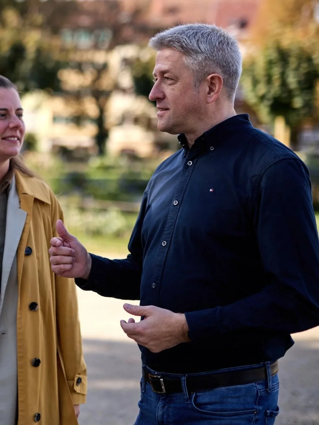Ein Mann im schwarzen Hemd spricht mit einer Frau im gelben Mantel im Freien bei Sonnenuntergang.