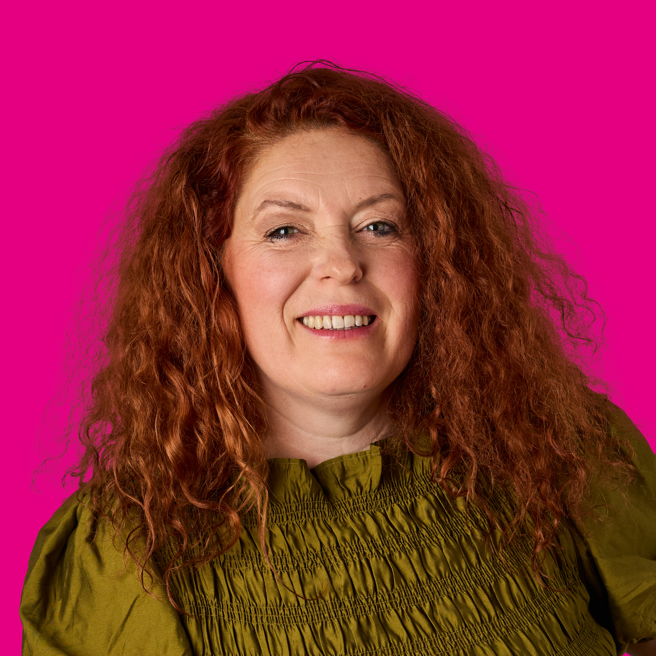 Porträt einer lächelnden Frau mit roten, lockigen Haaren vor rosa Hintergrund, trägt eine grüne Bluse.