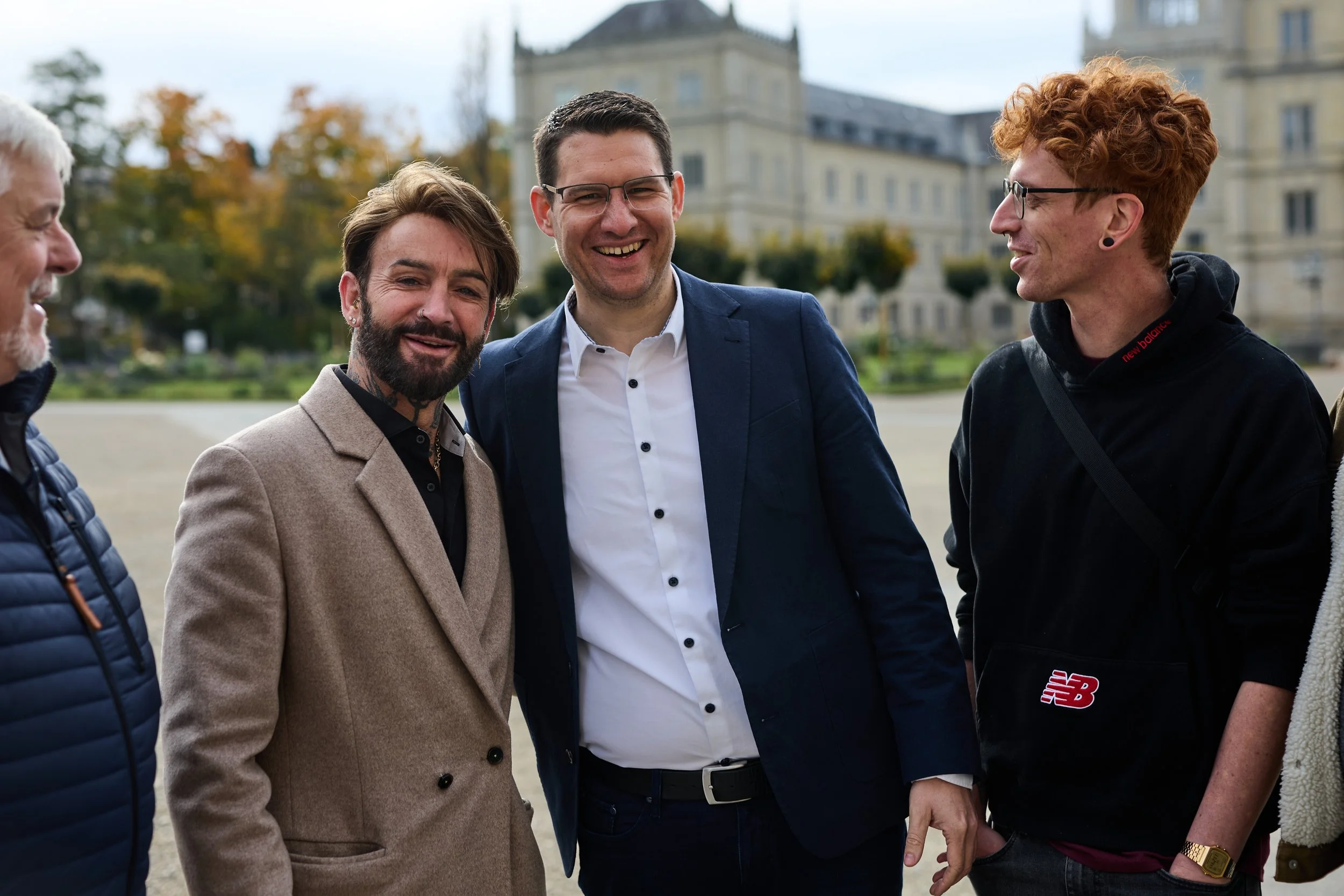 Fünf Männer im Gespräch, lächelnd, im Freien vor einem historischen Gebäude, mit Herbstbäumen im Hintergrund.