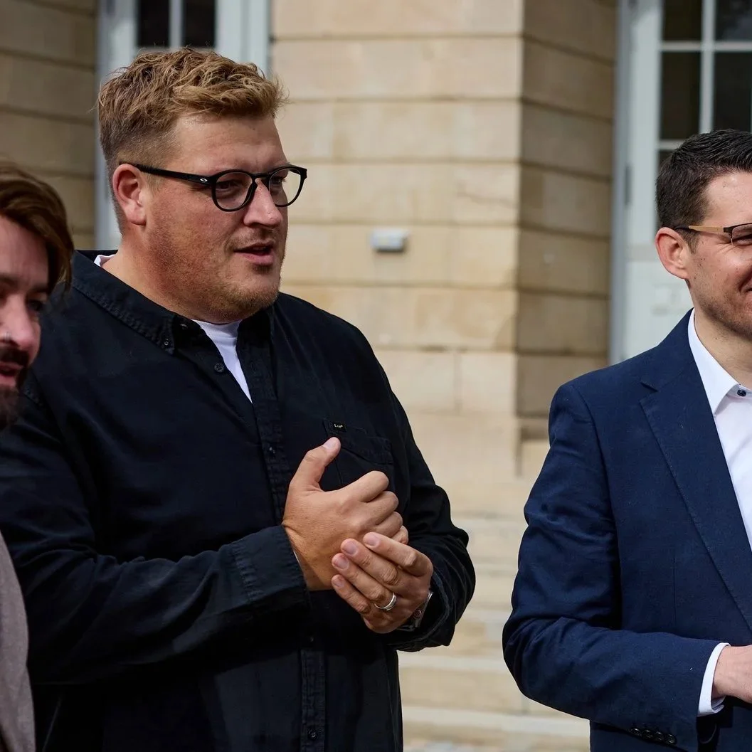 Drei Männer stehen draußen vor einem Gebäude, der Mann in der Mitte trägt eine schwarze Jacke und eine Brille, seine Hände sind vor der Brust verschränkt.