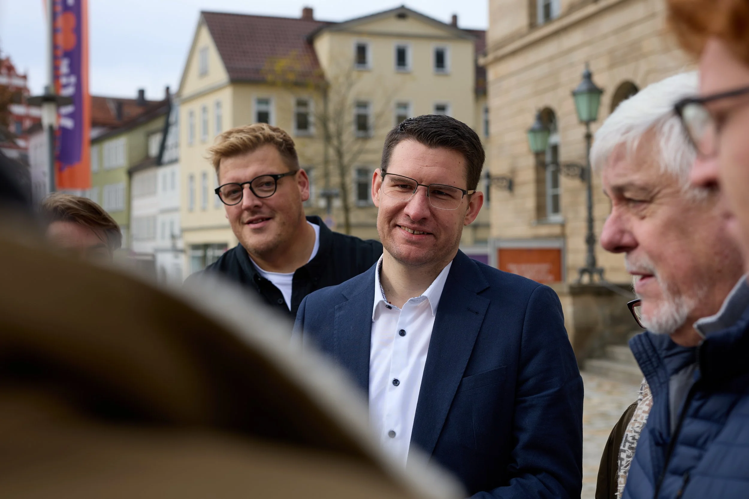 Eine Gruppe von Menschen im Gespräch auf einer Straße in einer Stadt mit alten Gebäuden, einige tragen Brillen.