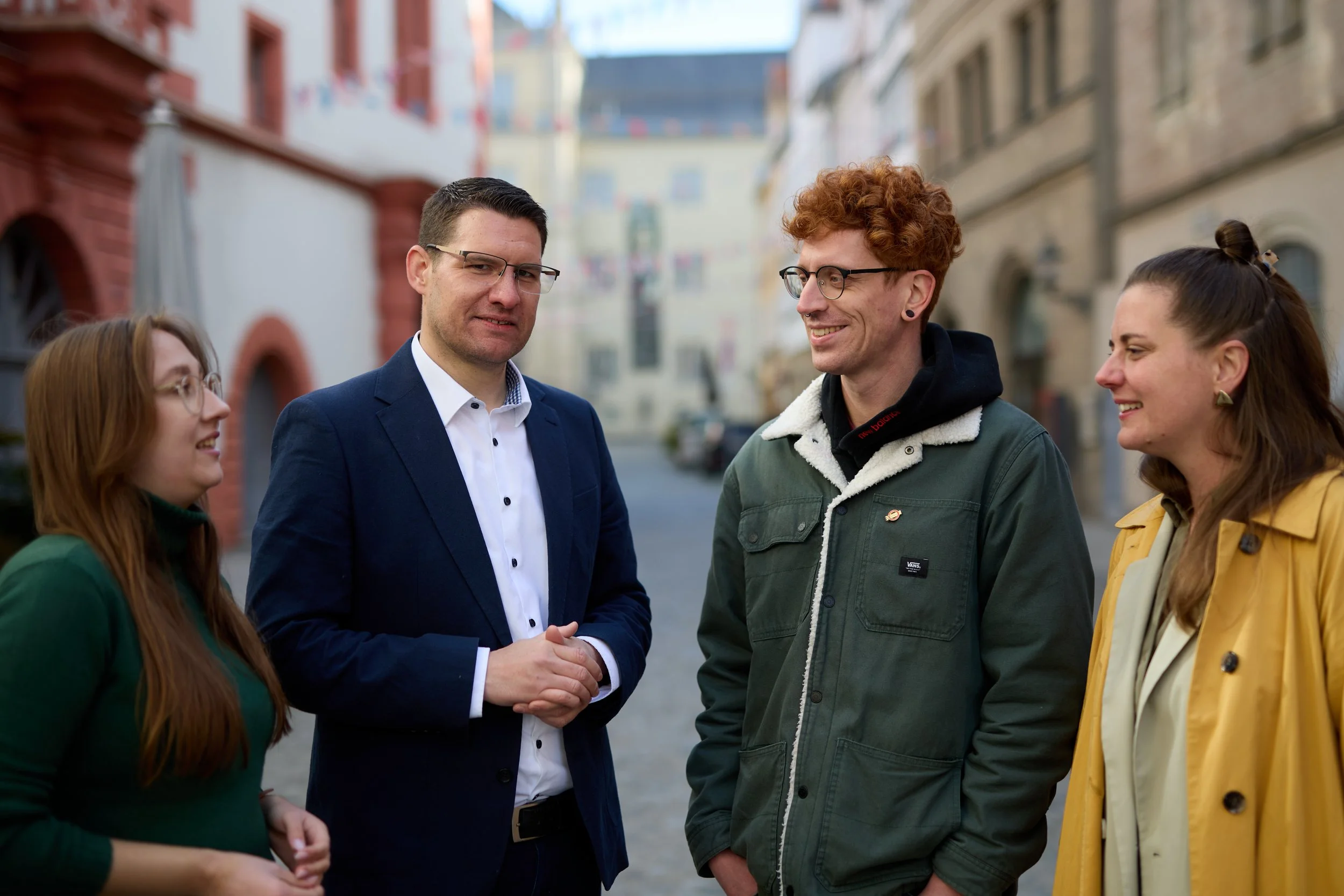 Vier Menschen unterhalten sich auf einer Straße in einer Stadt mit historischen Gebäuden im Hintergrund.