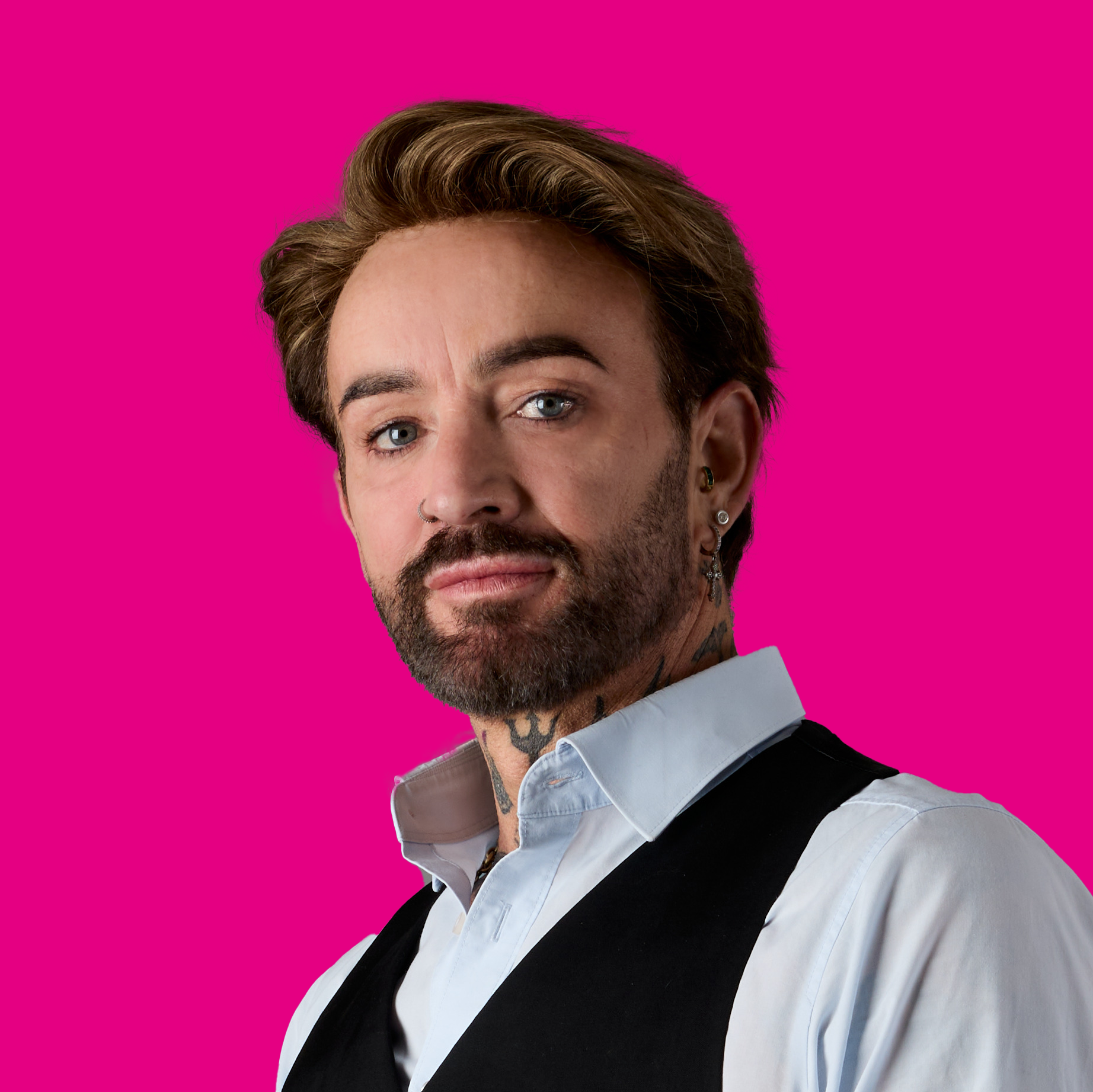 Porträt eines Mannes mit Tattoos, Piercings und braunem Haar vor einem pinken Hintergrund.