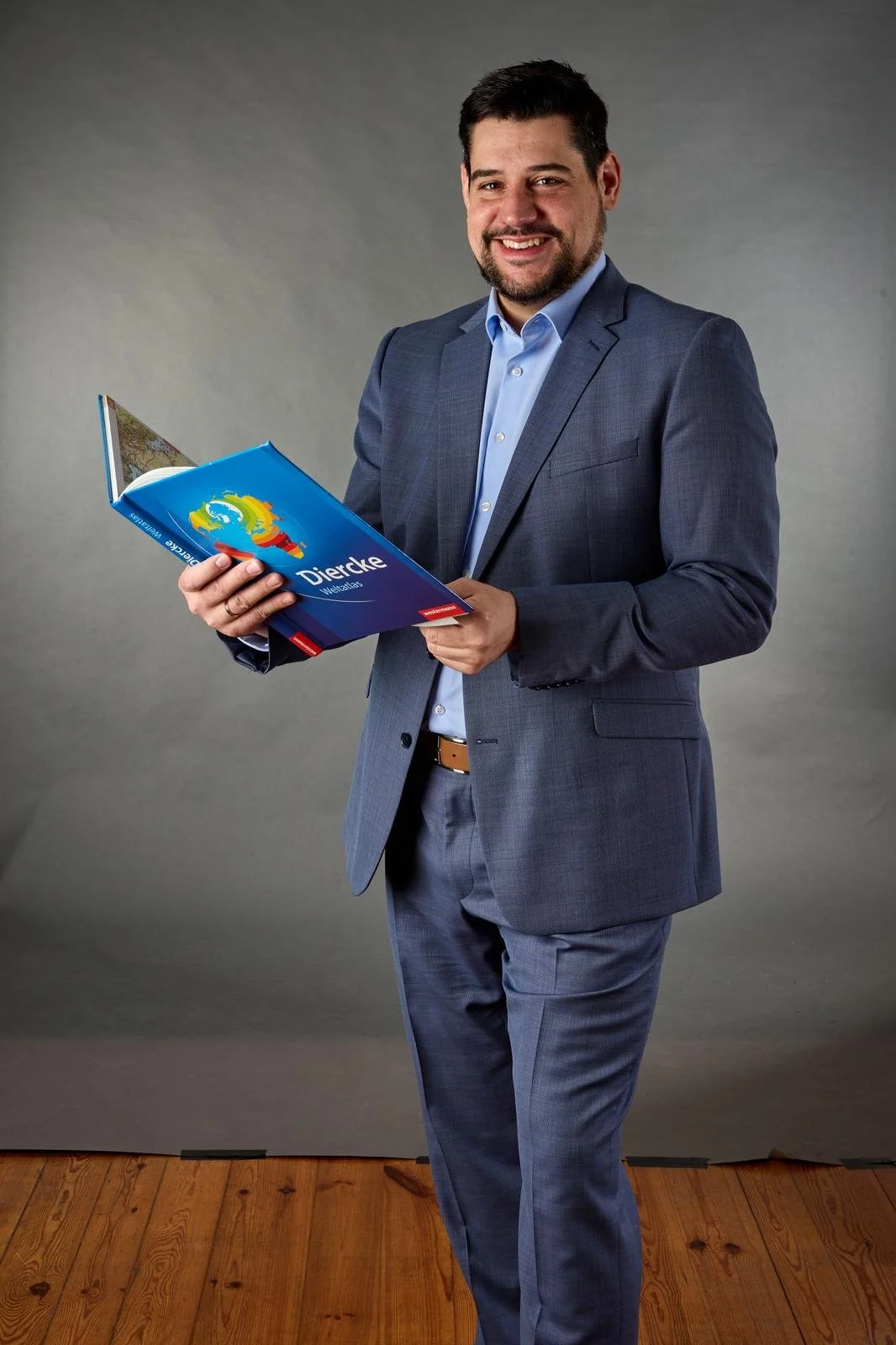 Ein Mann inBusiness attire hält einen Reiseführer für die Region Diercke Weltatlas, steht vor grauem Hintergrund und lächelt in die Kamera.