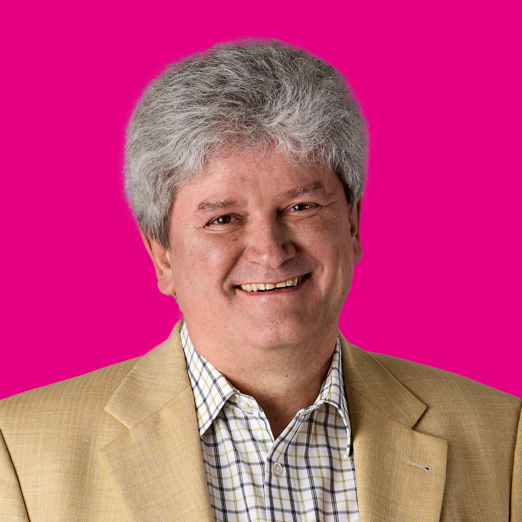Ein Mann mit grauem, leicht lockigem Haar, trägt einen gelben Anzug und ein kariertes Hemd, lächelt vor pinkem Hintergrund.
