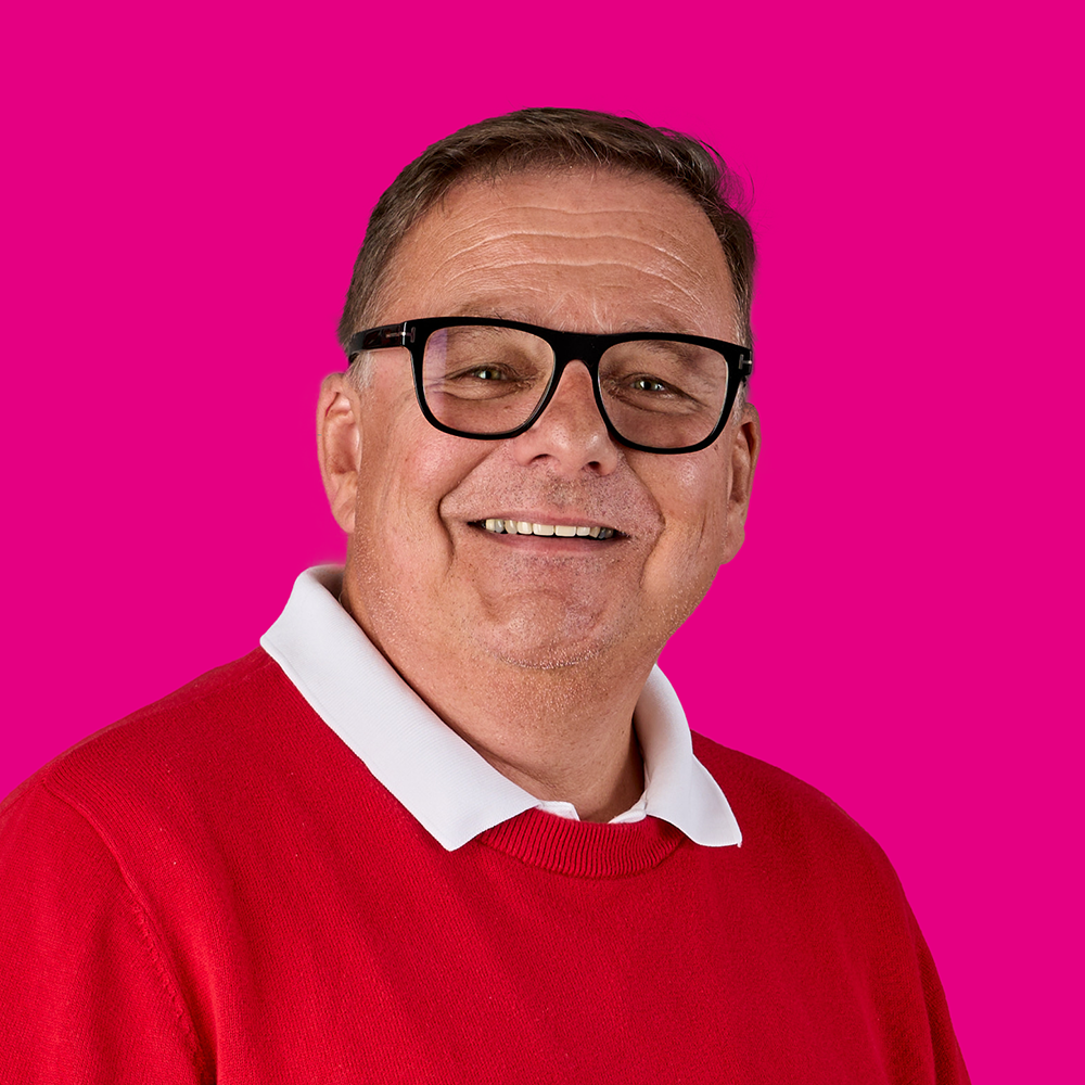 Lächelnder Mann mit Brille, rotes Pullover, weißes Hemd, auf pinkem Hintergrund.