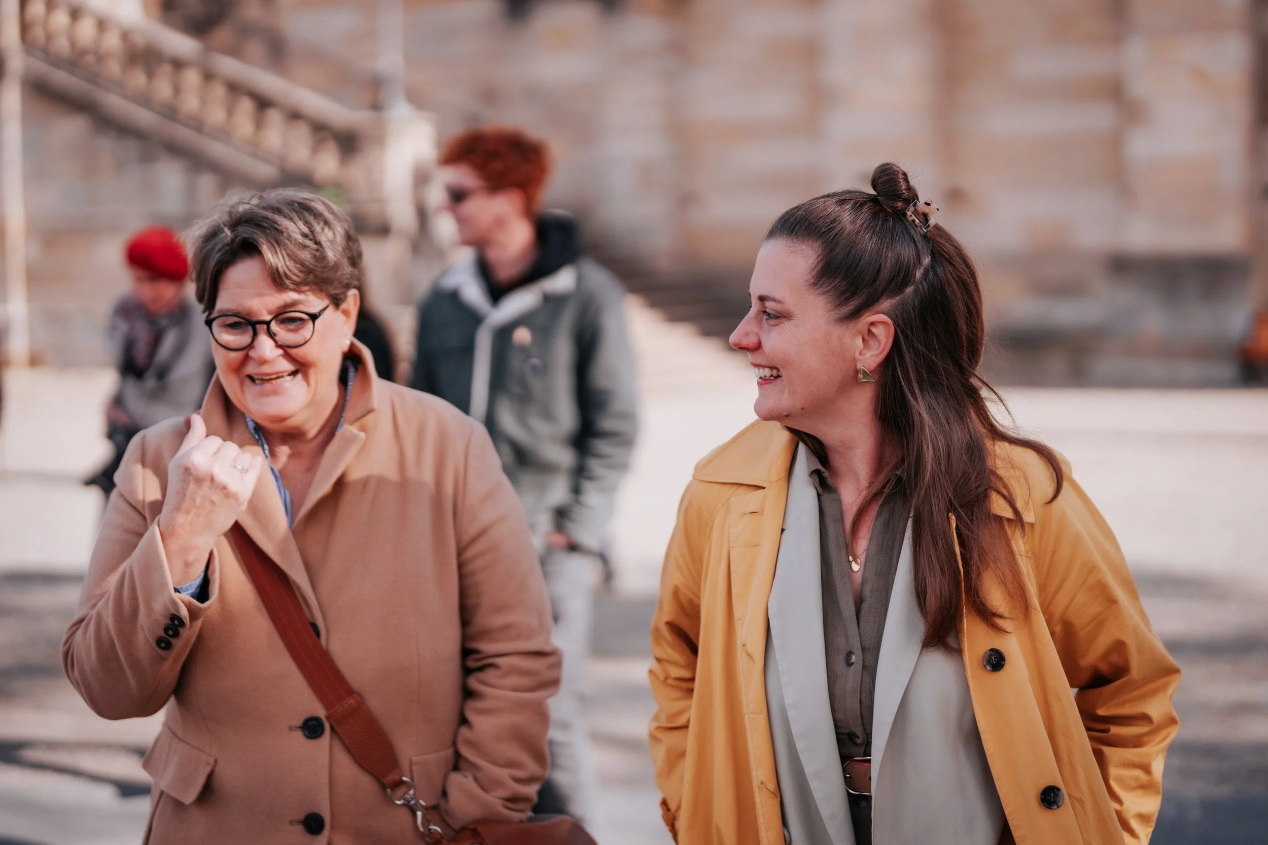 Zwei lachende Frauen im Gespräch, im Hintergrund eine weitere Person und Treppen.