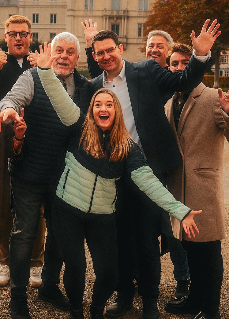 Eine Gruppe fröhlicher Menschen, hauptsächlich Männer, die draußen vor einem historischen Gebäude stehen und in die Kamera lächeln, während sie enthusiastisch winken.