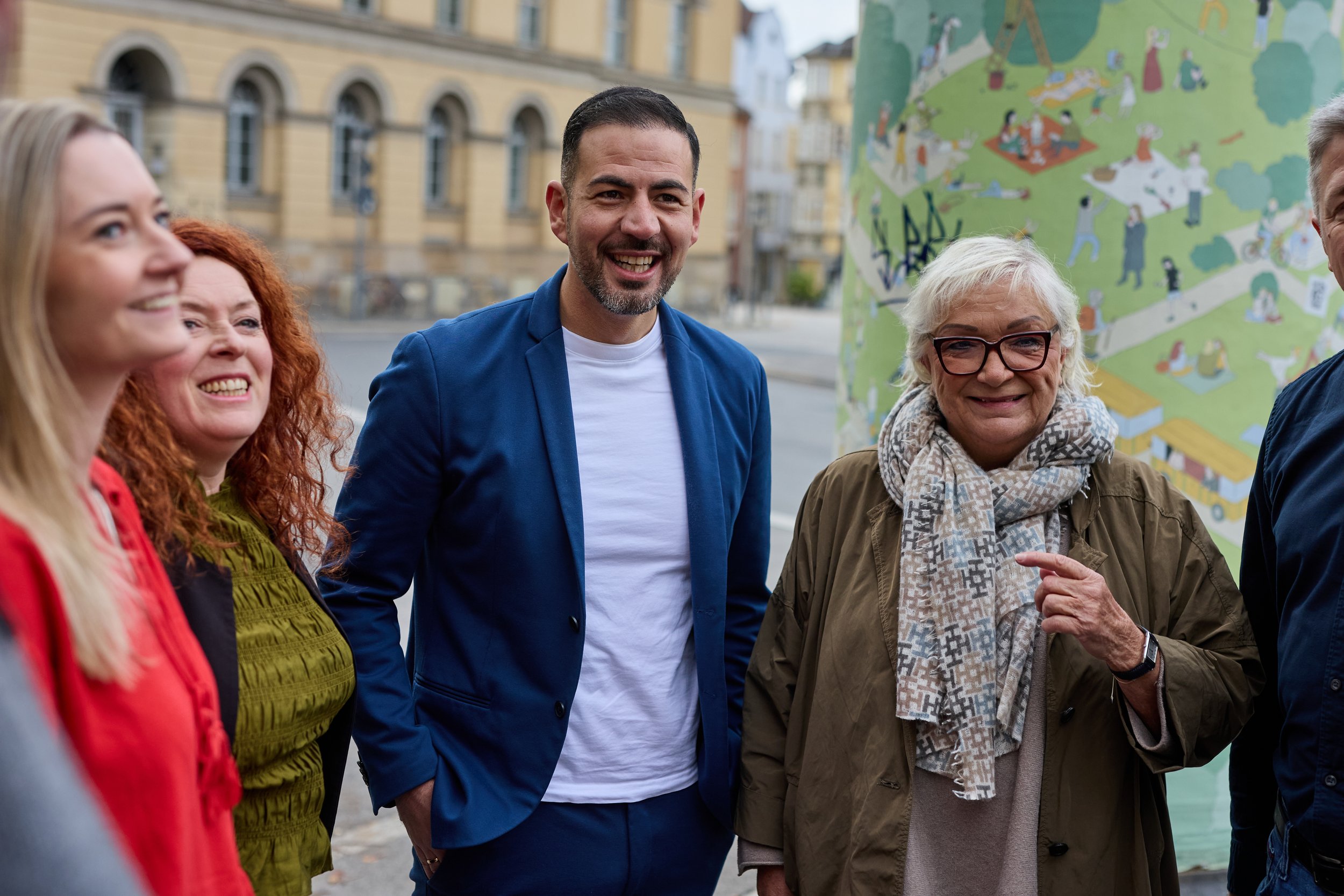 Gruppe von fünf fröhlichen Menschen, die draußen vor einer Wand mit buntem Wandbild stehen, lachend und sprechen