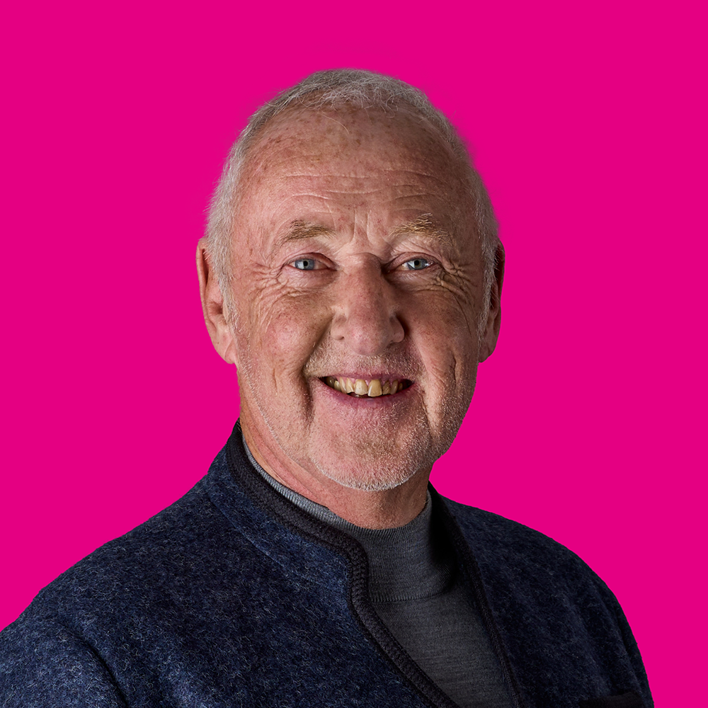 Lächelnder älterer Mann mit grauem Haar und grauem Bart vor pinkem Hintergrund.