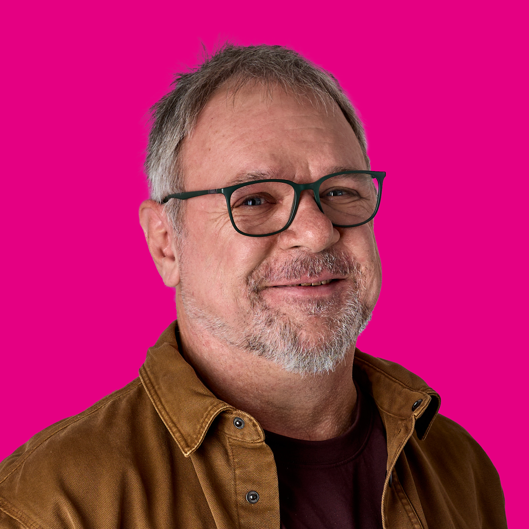 Ein lächelnder Mann mit Brille und grauem Bart vor pinkem Hintergrund.