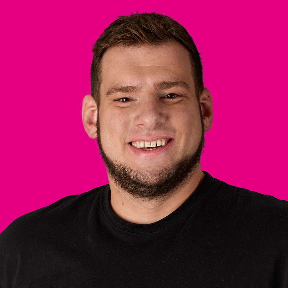 Ein lachender Mann mit braunen Haaren, Vollbart und schwarzen T-Shirt, vor pinkem Hintergrund.