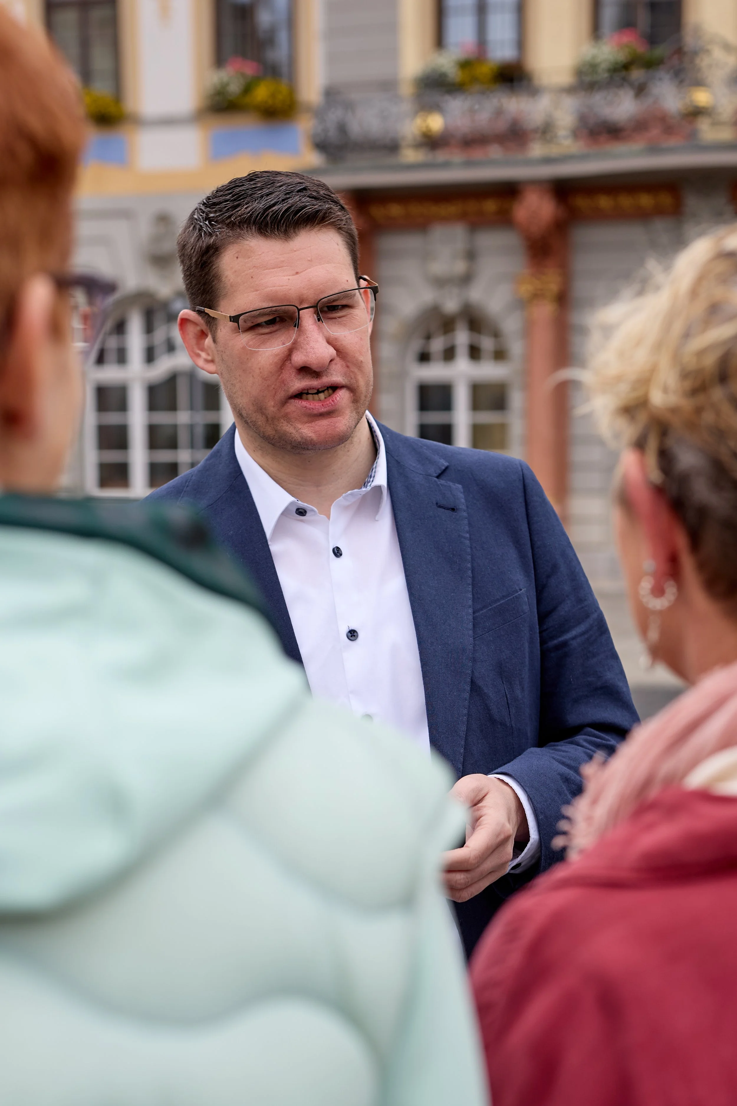 Ein Mann in einem blauen Anzug und mit Brille spricht mit drei Personen im Freien vor einem historischen Gebäude.