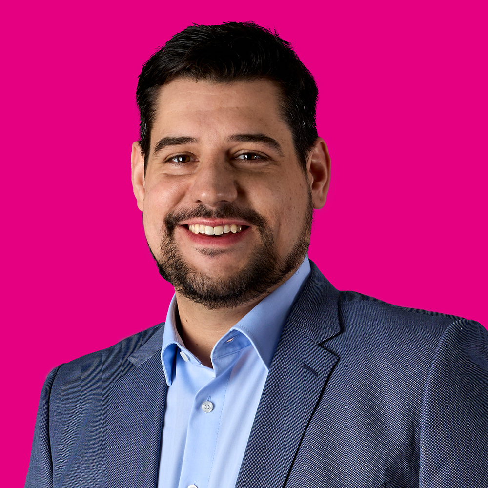 Ein lachender Mann in einem blauen Anzug und Hemd vor einem pinken Hintergrund.