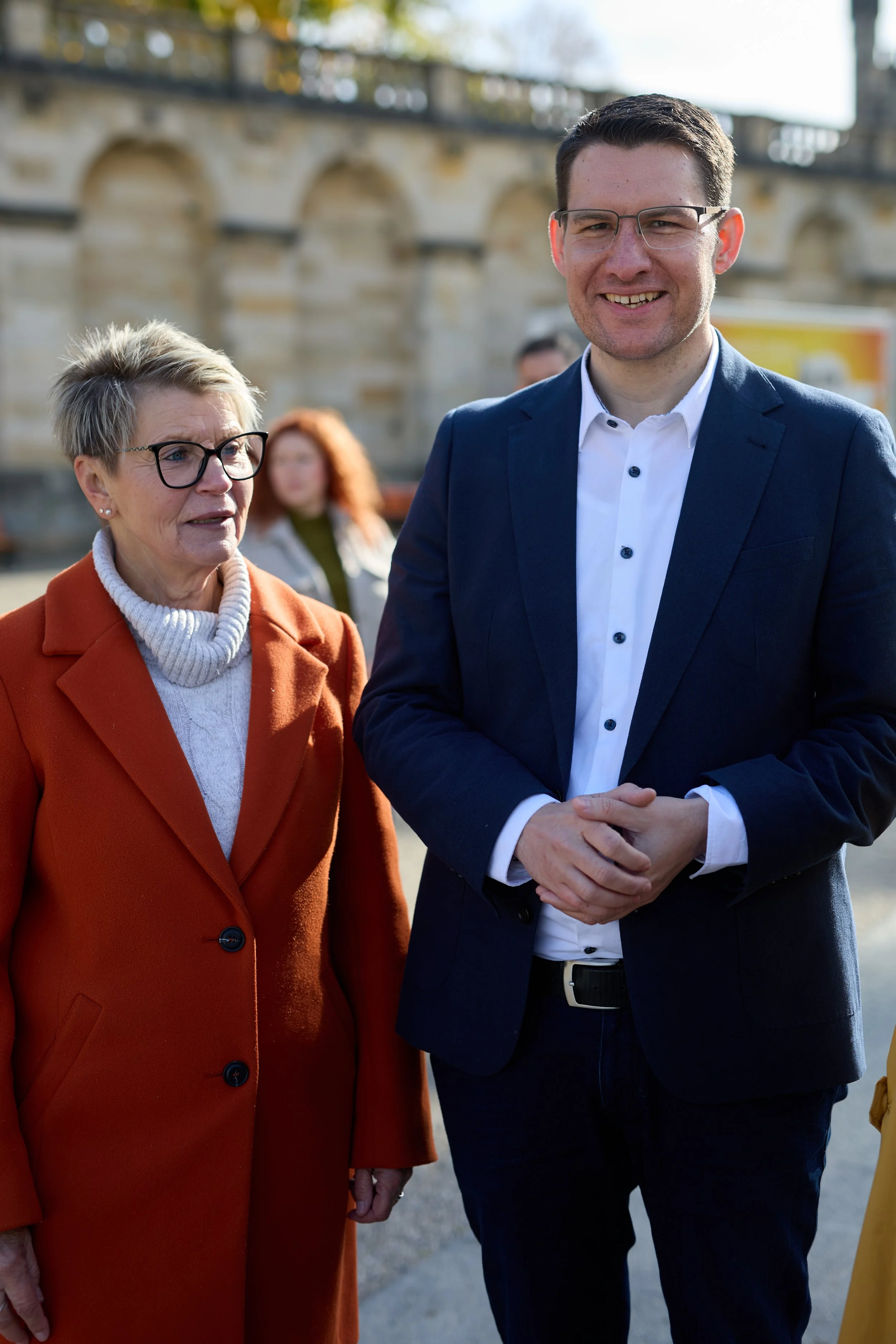 Ein Mann und eine Frau stehen im Freien vor einer Steinbrücke. Der Mann trägt einen dunklen Anzug und eine weiße Hemd, die Frau trägt einen orangefarbenen Mantel und eine graue Strickrolle. Im Hintergrund sind weitere Personen und ein gelbes Fahrzeug