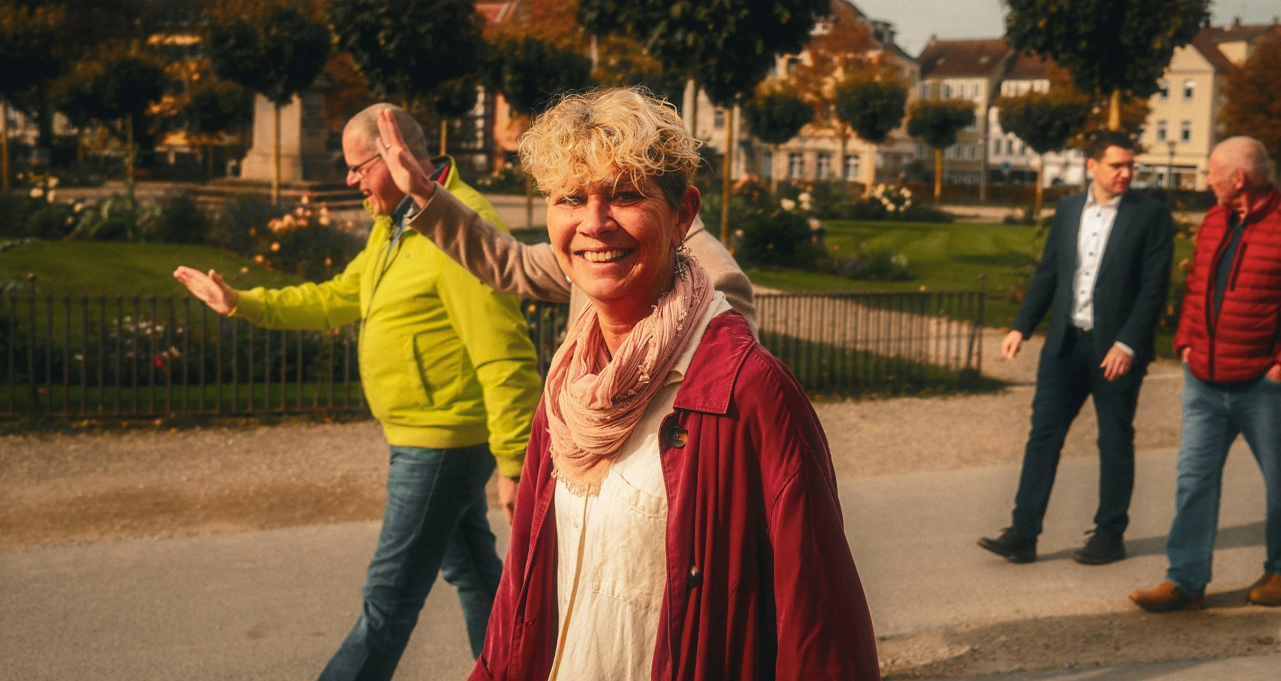 Eine fröhliche ältere Frau mit blond, lockigem Haar trägt eine rote Jacke und ein rosafarbenes Tuch, während sie auf einer Fußgängerbrücke in einer parkähnlichen Umgebung unterwegs ist, umgeben von anderen Menschen im Gespräch.
