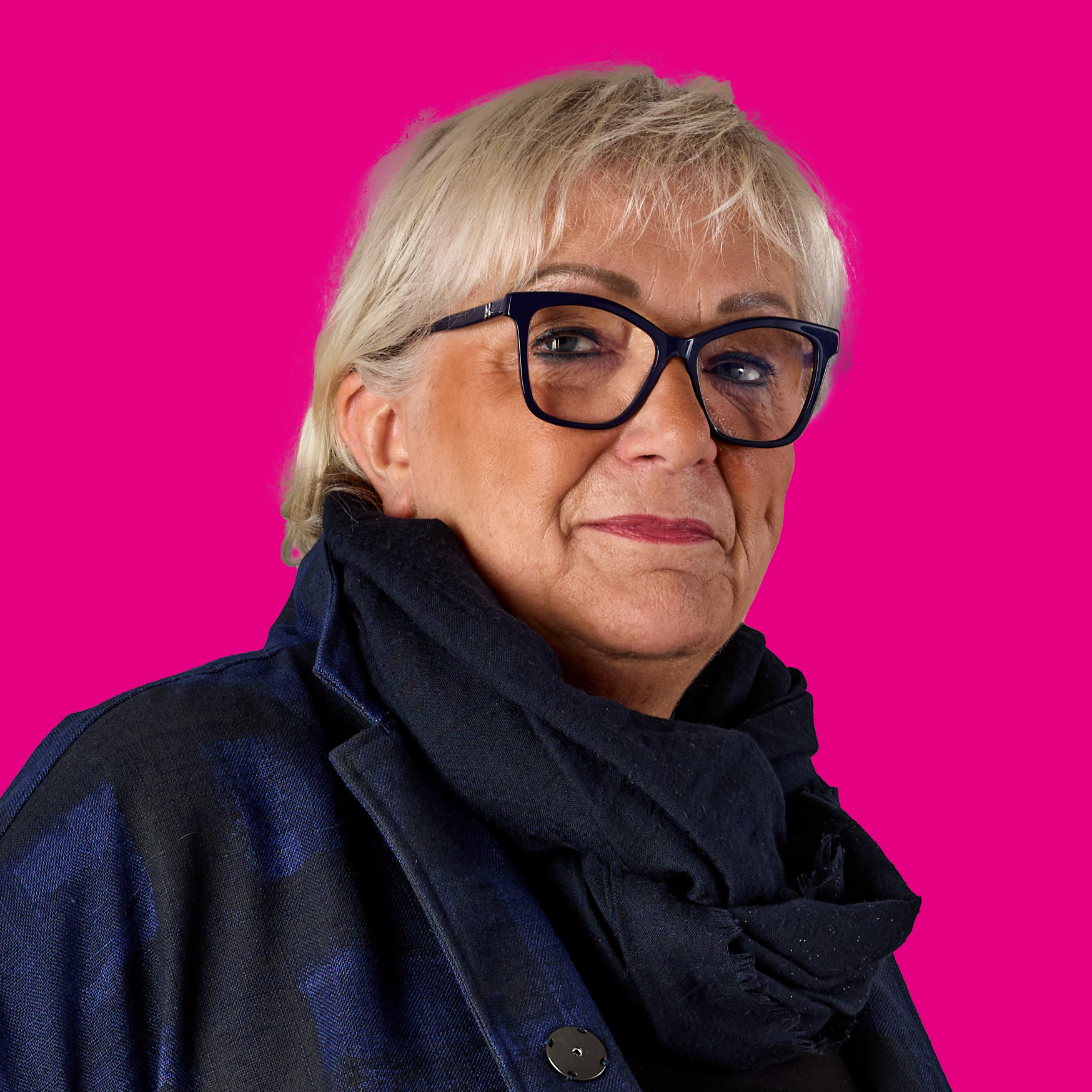 Ein älterer, ernst blickender Mensch mit kurzen, grauen Haaren, schwarzen Brillen und schwarzem Kleid vor einem pinken Hintergrund.