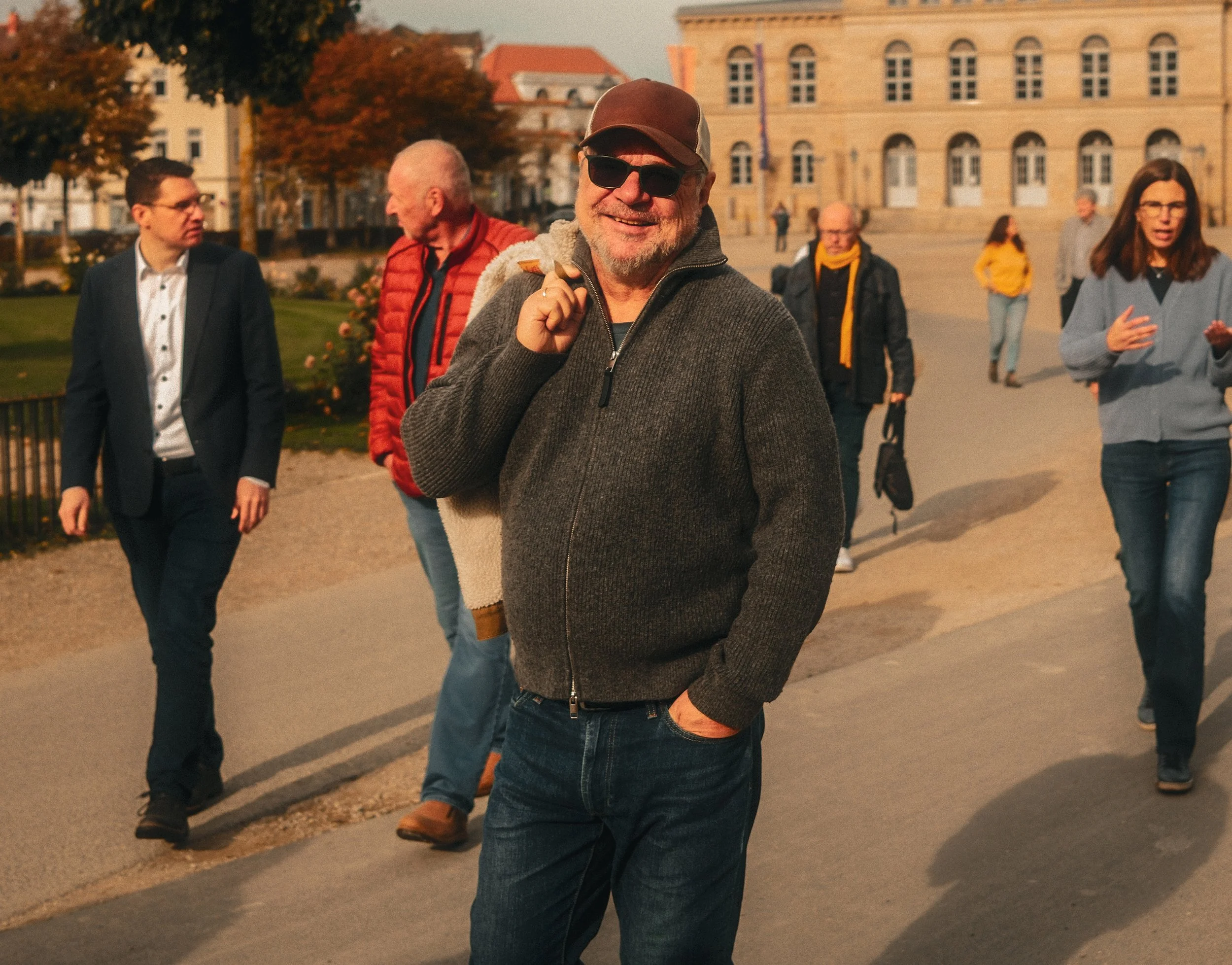 Ein Mann trägt eine Sonnenbrille, einen braunen Hut und einen grauen Pullover, während er in einer Gruppe von Menschen in einem Stadtpark unterwegs ist, im Hintergrund Gebäude und Bäume mit Herbstlaub.