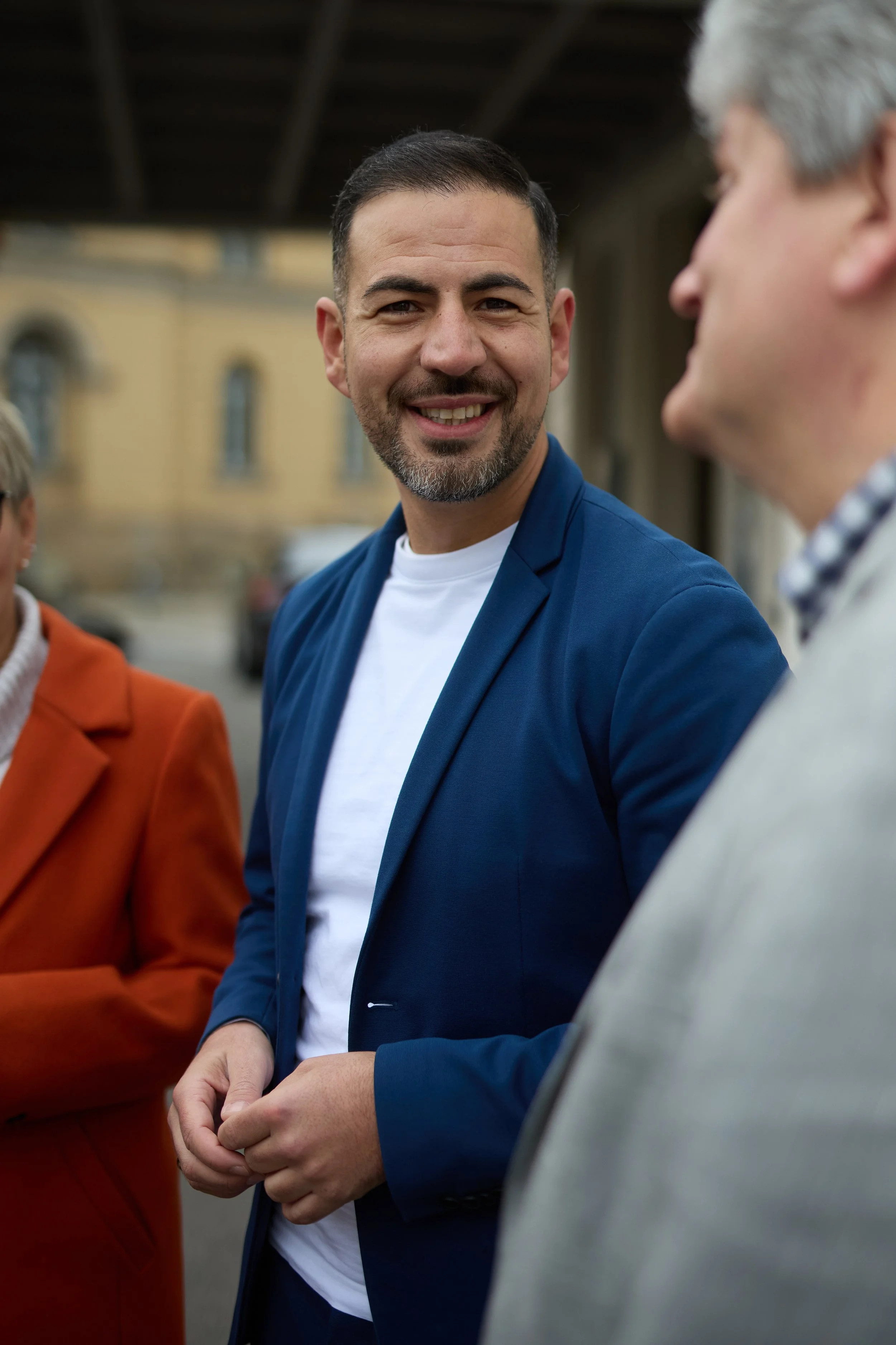 Ein Mann im blauen Anzug, lächelnd, im Gespräch mit zwei anderen Personen bei einem Outdoor-Event.