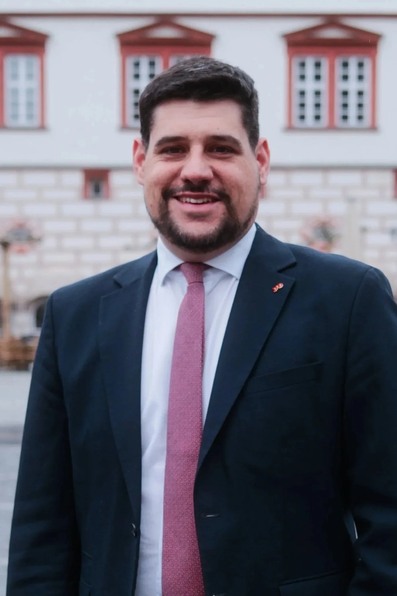 Ein lachender Mann in einem blauen Anzug und Hemd vor einem pinken Hintergrund.
