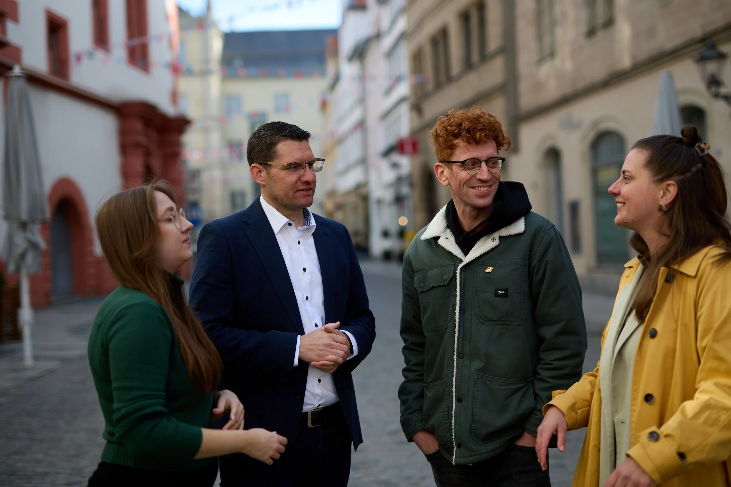 Vier Menschen stehen auf einer Stadtstraße im Gespräch, im Hintergrund sind alte Gebäude und bunte Wimpel schwebend.