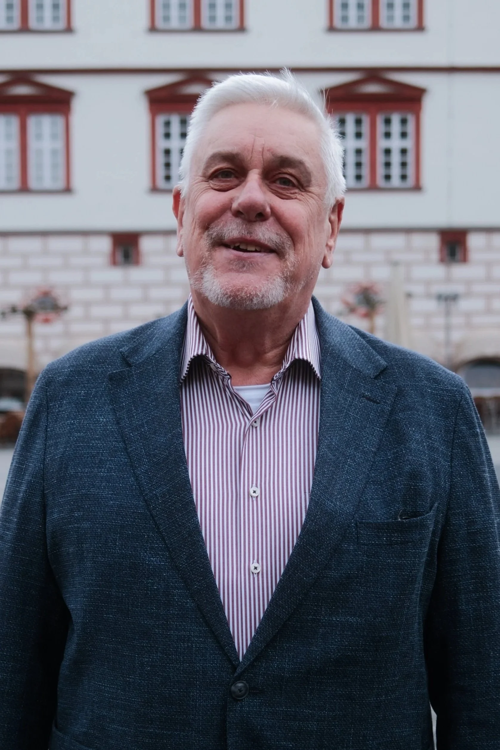 Porträt eines älteren Mannes mit weißen Haaren und Bart, trägt einen dunkelblauen Anzug und ein weißes Hemd, vor pinkem Hintergrund.