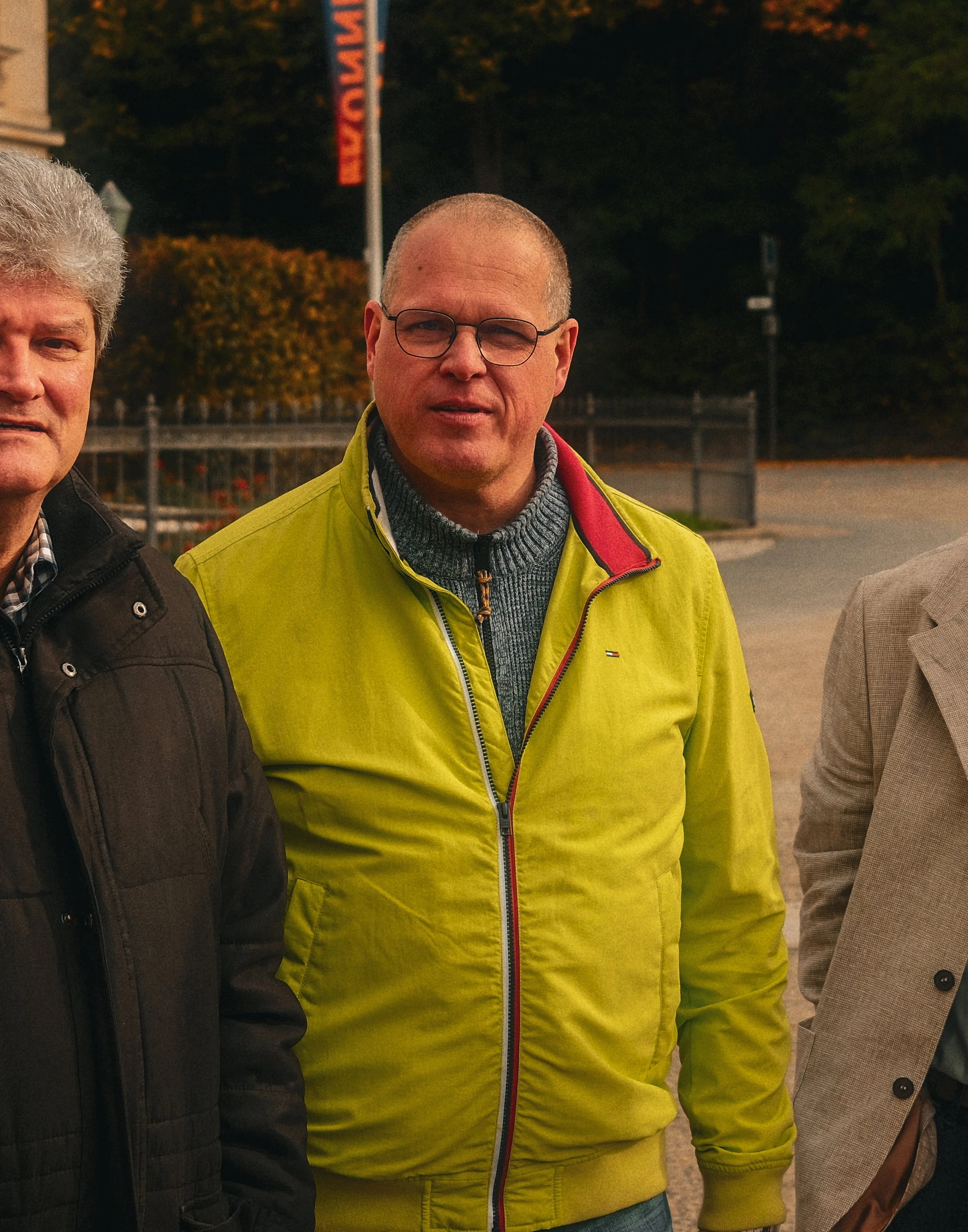 Drei Männer stehen im Freien, einer trägt eine gelbe Jacke, der andere eine schwarze Jacke, und der dritte eine beige Jacke, bei Dämmerung.