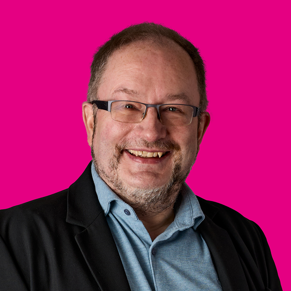 Porträt eines lachenden Mannes mit Brille vor pinkem Hintergrund.