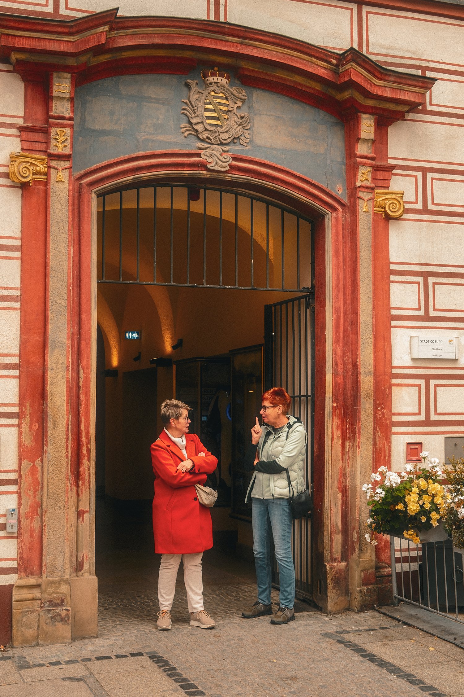 Zwei Frauen unterhalten sich vor einem Eingang zu einem historischen Gebäude mit roten und beige Steinen und einem Wappen oberhalb. Es sind Blumentöpfe mit weißen und gelben Blumen neben dem Eingang zu sehen.