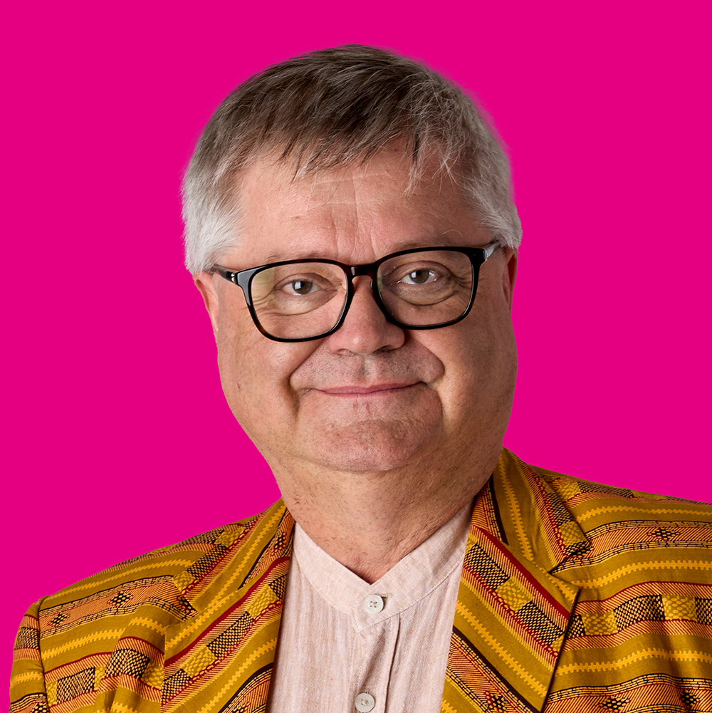 Porträt eines Mannes mit grauem Haar, Brille, gelber und bunter Jacke vor rosa Hintergrund.
