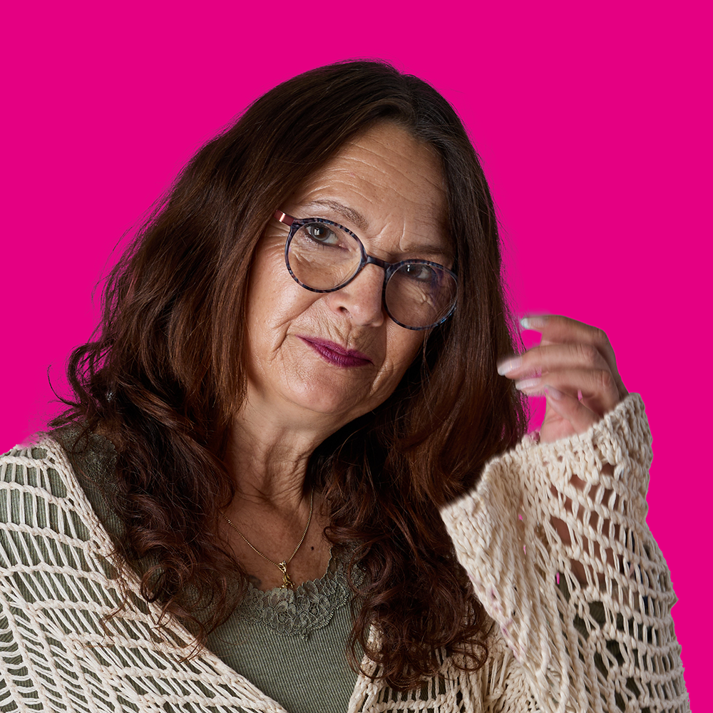Porträt einer ernst aussehenden älteren Frau mit Brille und braunen Locken vor pinkem Hintergrund.