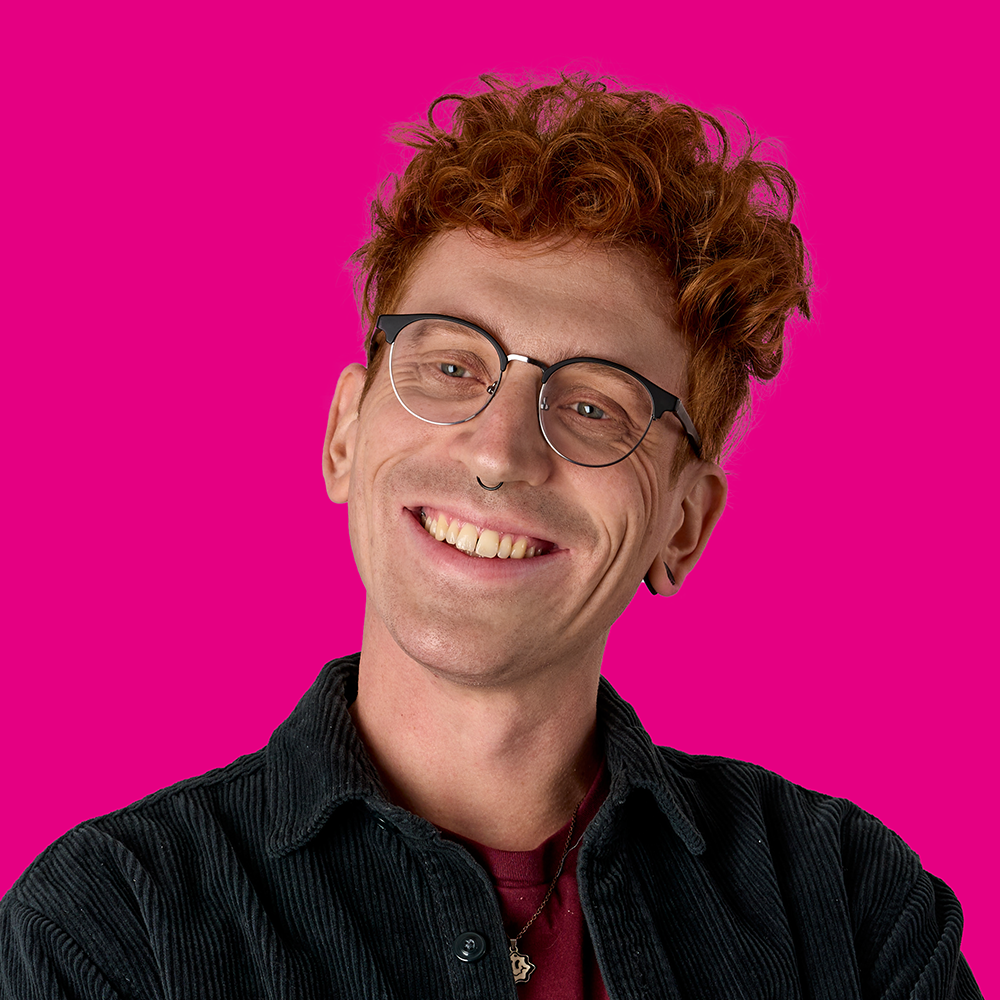 Porträt eines lachenden Mannes mit roten, lockigen Haaren, Brille, Piercings und dunklem Shirt vor pinkem Hintergrund