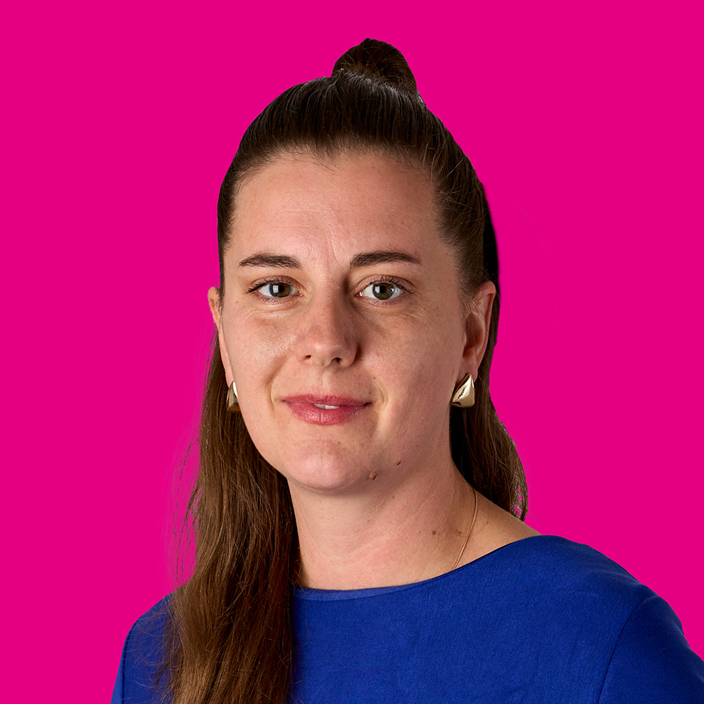 Porträt einer Frau mit braunen Haaren, die in eine Kamera blickt, trägt eine blaue Bluse, vor pinkem Hintergrund.