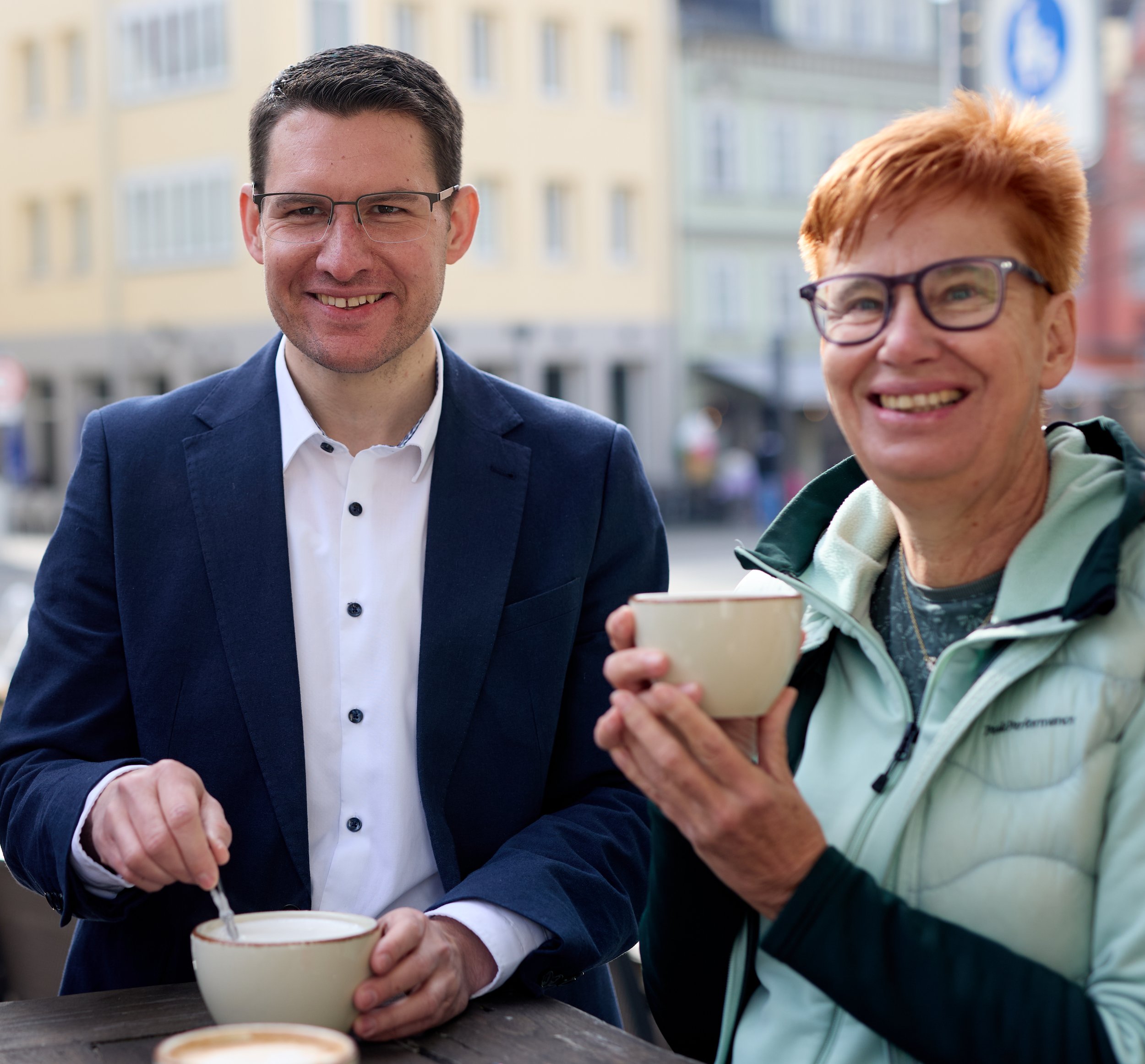 Zwei lachende Personen, ein Mann im Anzug und eine Frau mit Brille und Taschenlampe, trinken Kaffee im Freien auf einer Stadtstraße.