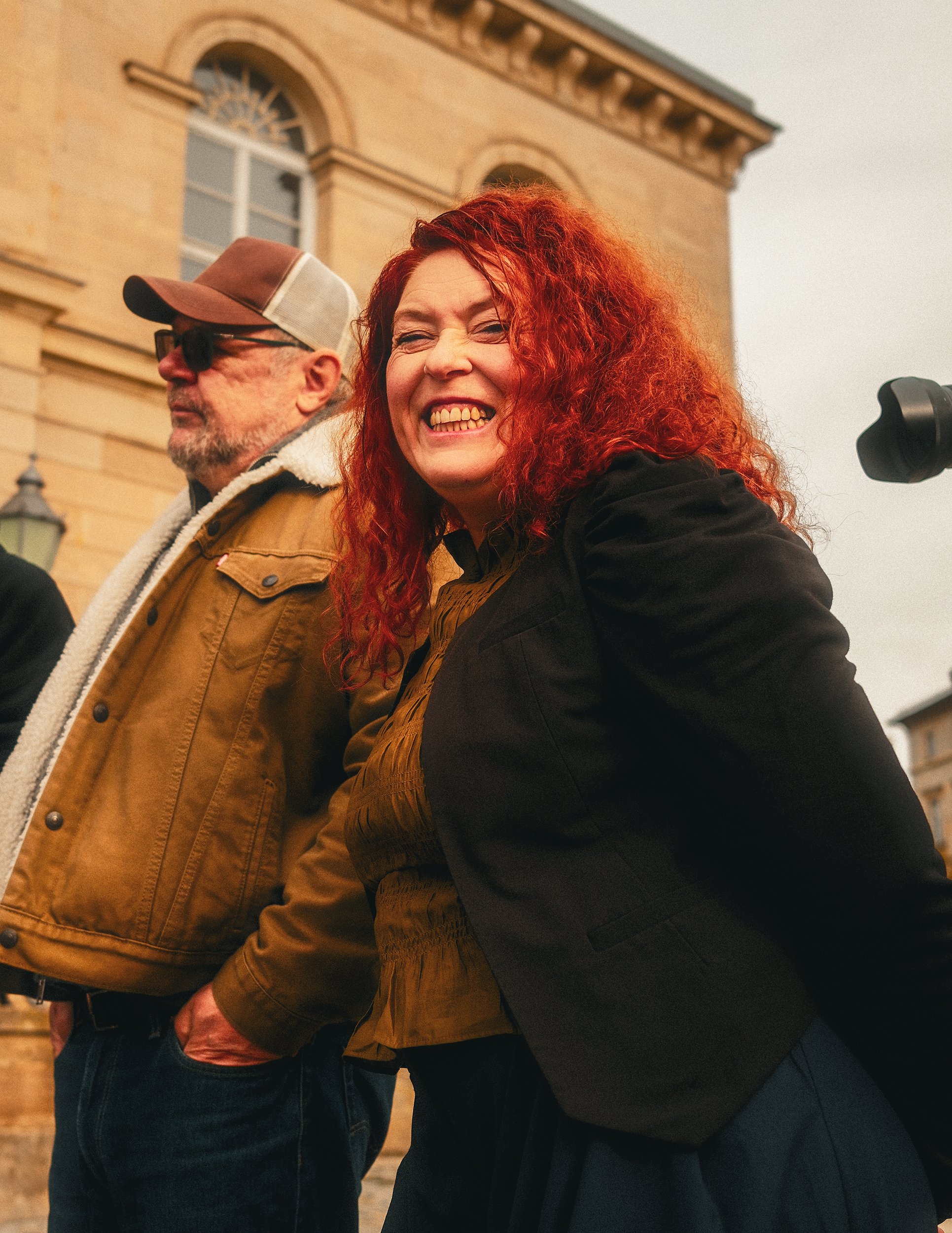 Eine lachende Frau mit roten, lockigen Haaren steht neben einem Mann mit Brille und Baseballkappe vor einem historischen Gebäude.