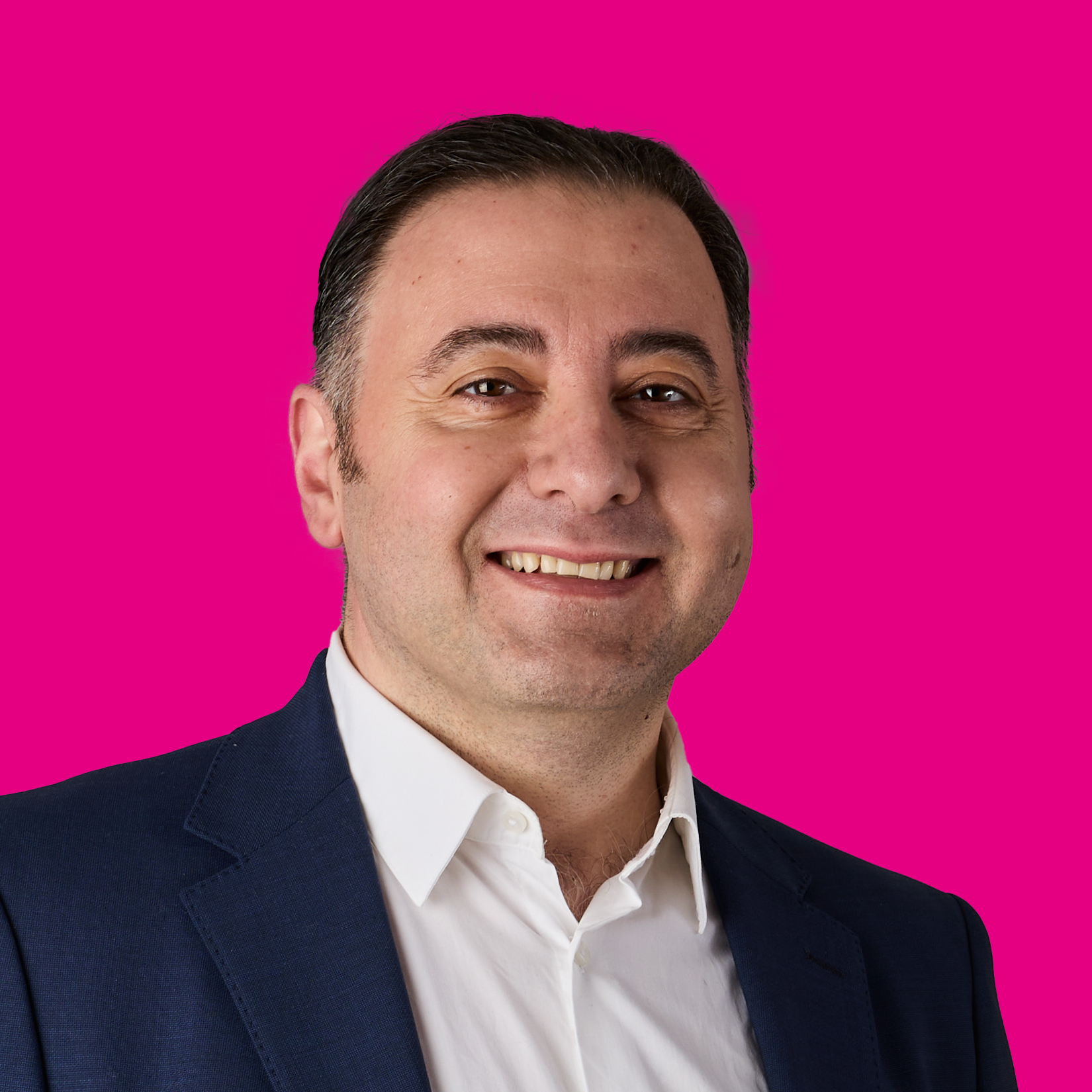 Porträt eines lachenden Mannes in formeller Kleidung vor pinkem Hintergrund.