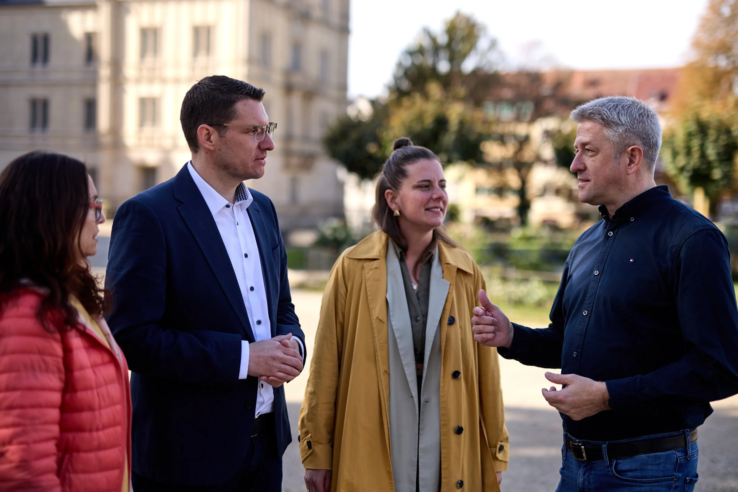 Vier Menschen im Gespräch im Freien, drei Frauen und ein Mann, der eine redet, während die anderen zuhören, im Hintergrund ein historisches Gebäude und Bäume, es ist herbstlich.